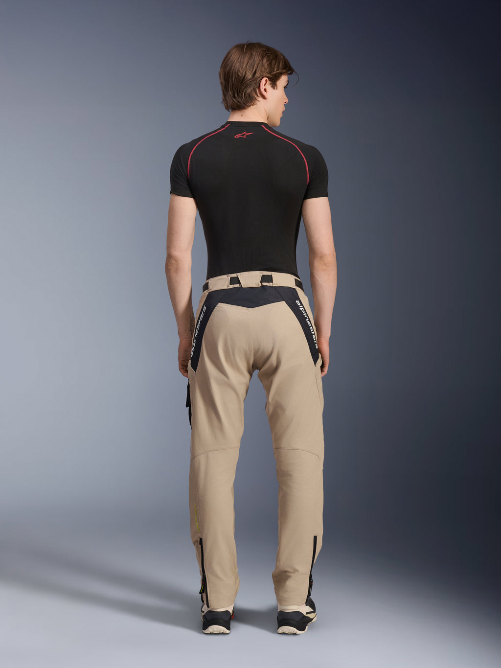 Alpinestars Acteon Pants in Sand, moderni pantaloni da moto urban visti dal retro, con pannelli neri e logo Alpinestars bianco, vestibilità affusolata con zip sul fondo, indossati con base layer nero a maniche corte e sneakers.