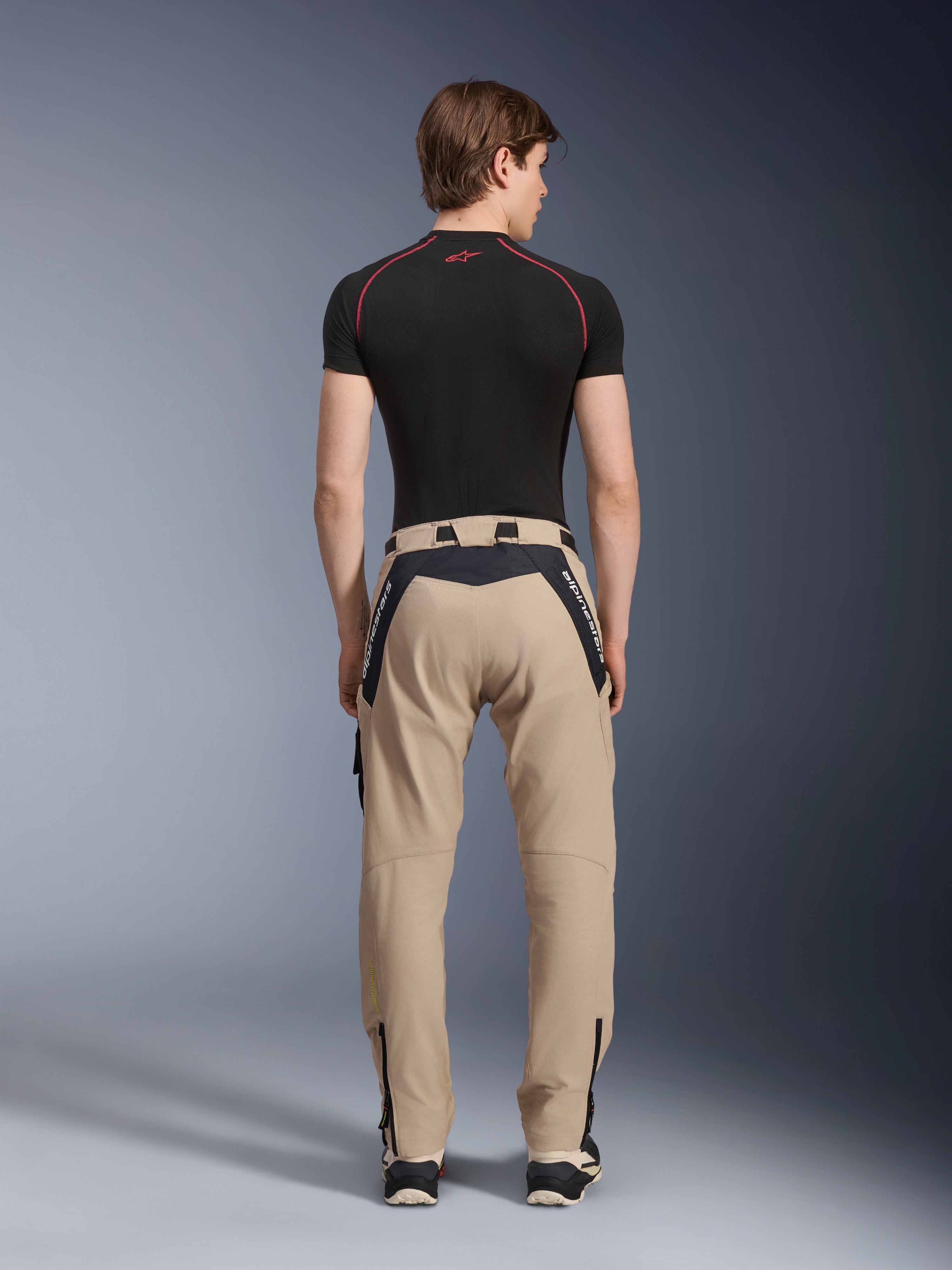Alpinestars Acteon Pants in Sand, moderni pantaloni da moto urban visti dal retro, con pannelli neri e logo Alpinestars bianco, vestibilità affusolata con zip sul fondo, indossati con base layer nero a maniche corte e sneakers.