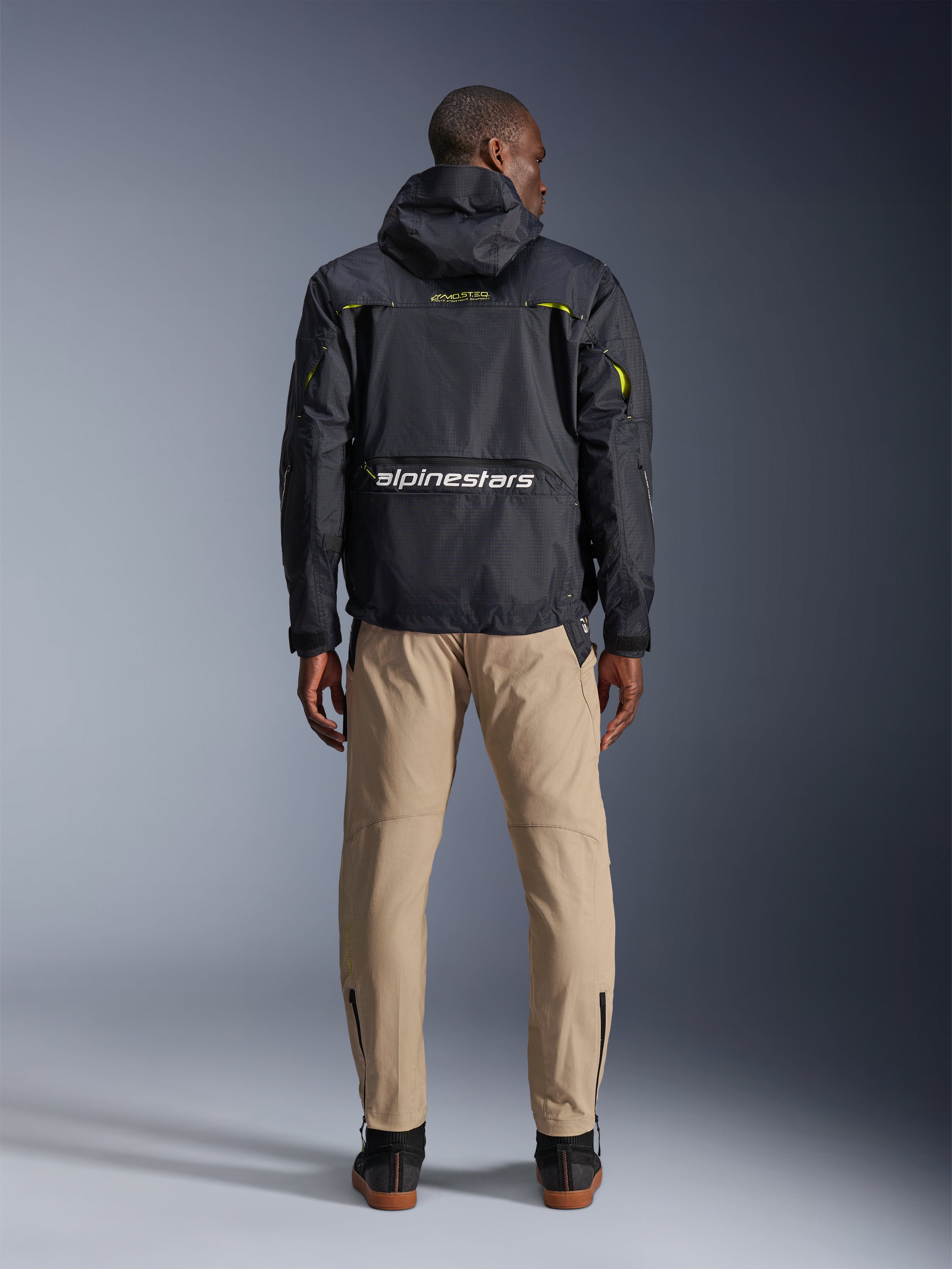 Pantaloni Alpinestars Acteon, pantaloni da moto urban, colore sabbia, visti dal retro su un modello che indossa una giacca tecnica nera con cappuccio e stivali scuri, caratterizzati da una gamba affusolata con zip alle caviglie e una silhouette pulita e moderna.