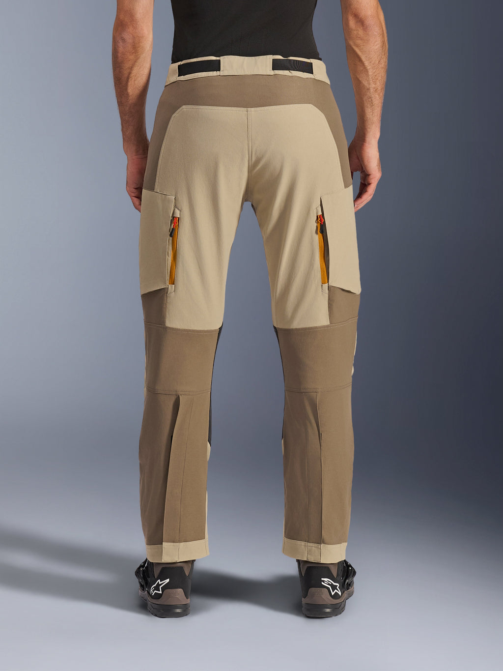 Alpinestars Maxdura Dual Pants, Walnut Aluminum, vista posteriore di pantaloni da moto adventure con pannelli color sabbia e toni della terra, prese d'aria con cerniere arancioni e design sopra lo stivale, indossati con stivali Alpinestars neri