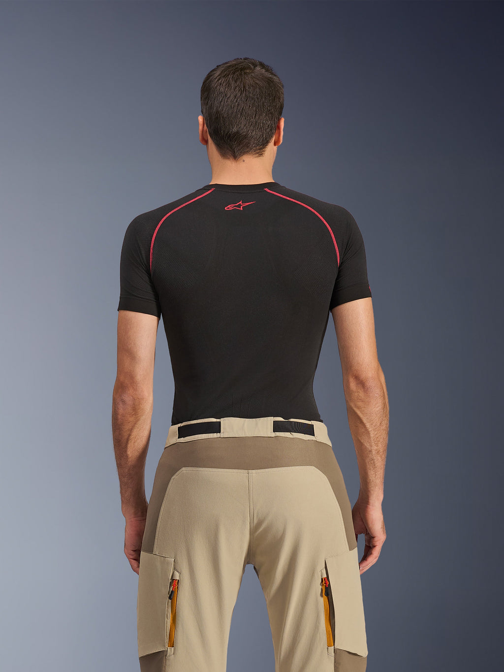 Alpinestars Maxdura Dual Pants, pantaloni da moto adventure, Walnut Aluminum, vista posteriore con pannello rinforzato sulla seduta e tessuto elasticizzato beige, tasche cargo con cerniera e tiretti arancioni, indossati con maglia base layer nera Alpinestars
