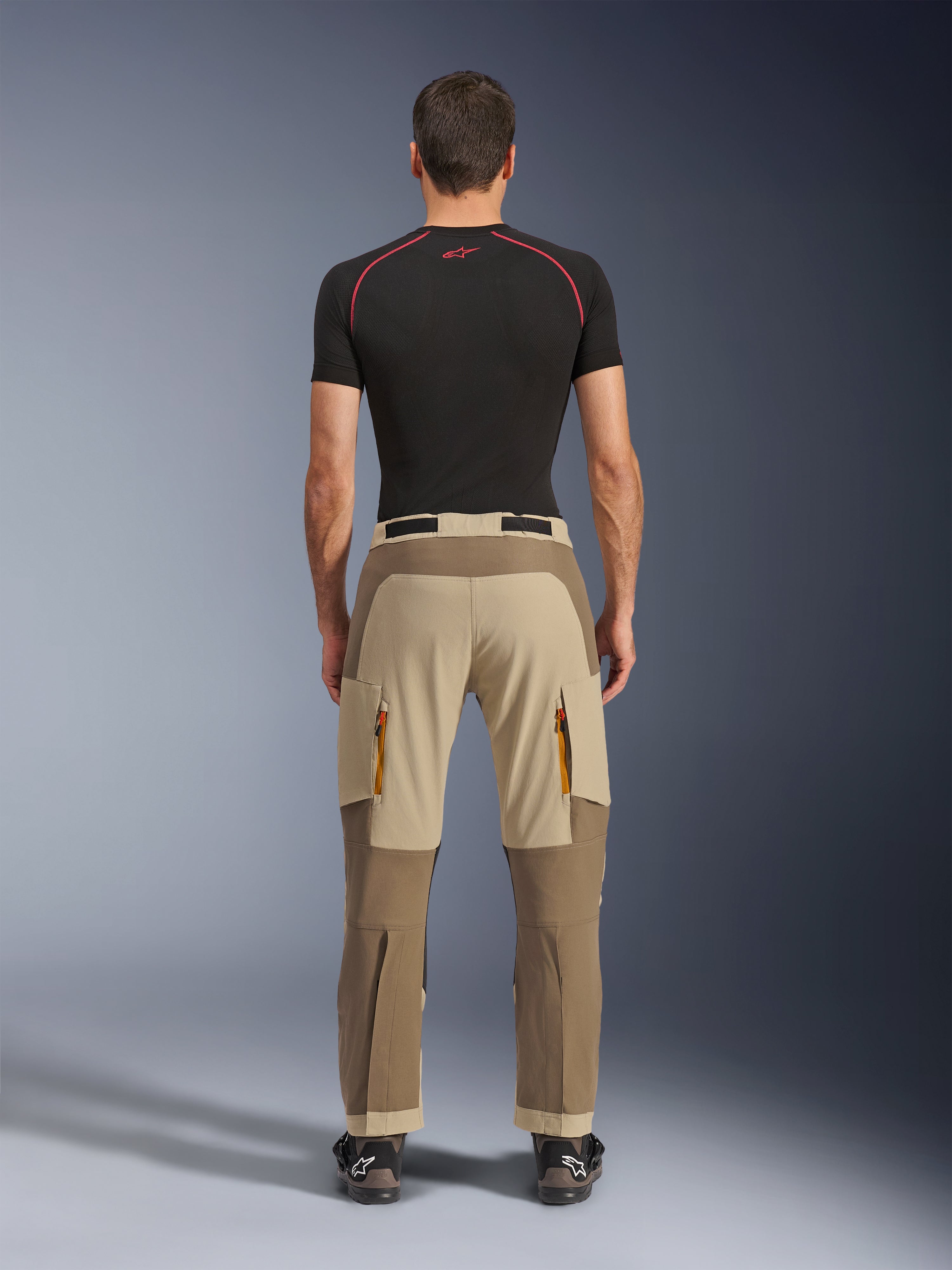 Alpinestars Maxdura Dual Pants in Walnut Aluminum, vista posteriore che mostra il design bicolore marrone e beige, pantaloni da moto adventure con tasche cargo con zip e seduta rinforzata, indossati con maglia a compressione nera e stivali da moto