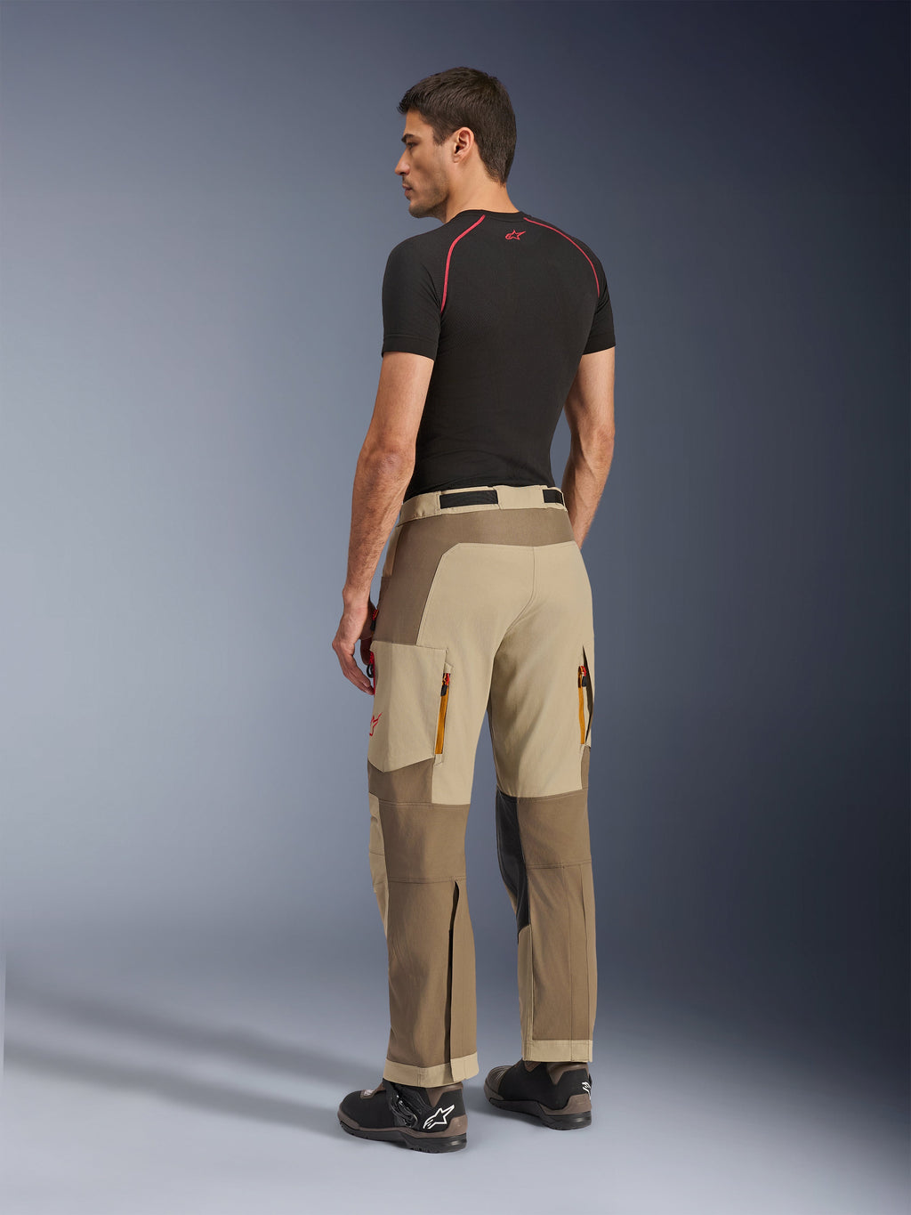 Alpinestars Maxdura Dual Pants, pantaloni da moto Adventure e Dual-Sport, Walnut Aluminum, vista posteriore indossata da un modello con base layer nero e stivali da moto, caratterizzati da costruzione multi-pannello con prese d'aria posteriori con zip e rinforzi.