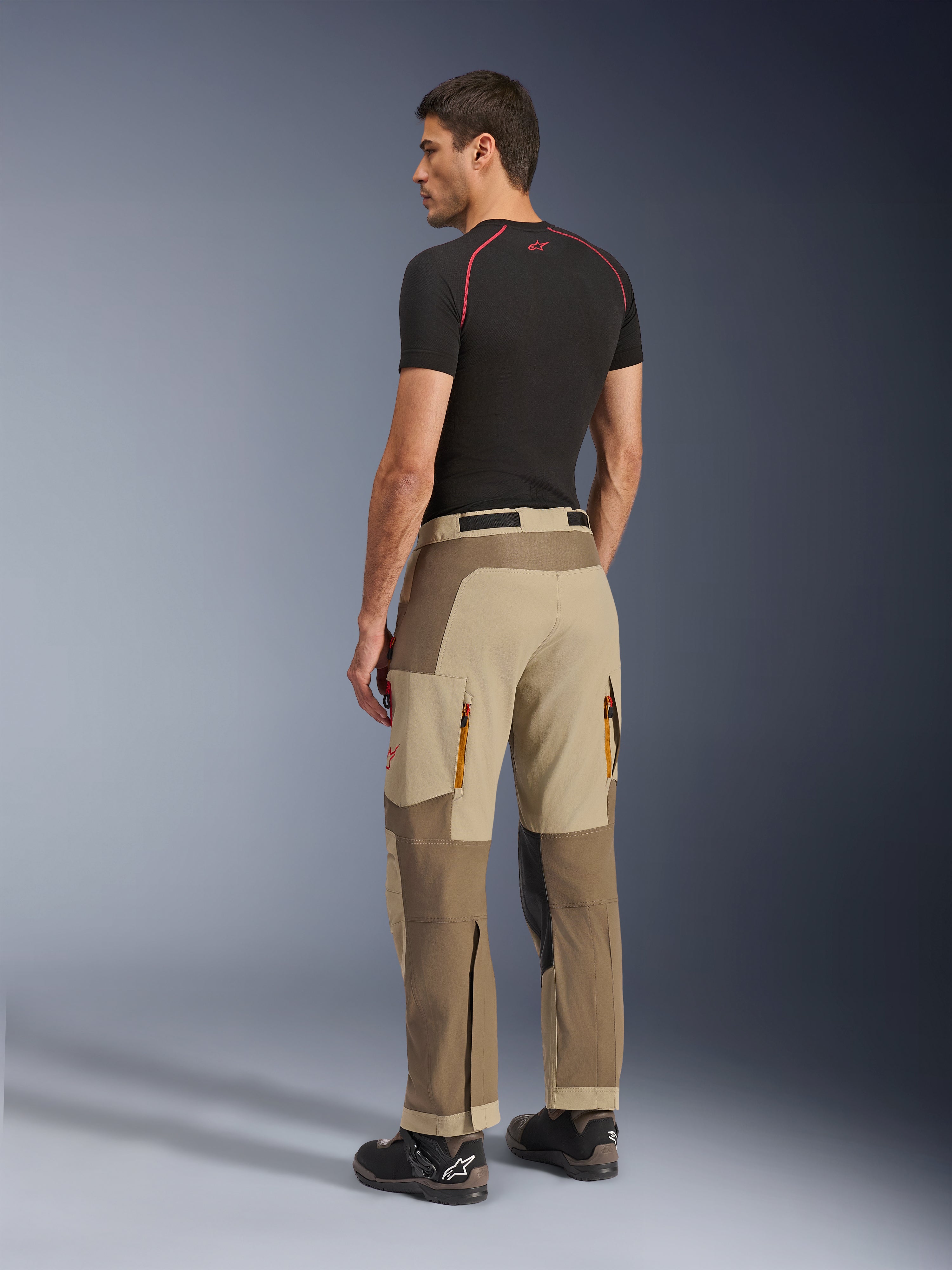Alpinestars Maxdura Dual Pants, pantaloni da moto Adventure e Dual-Sport, Walnut Aluminum, vista posteriore indossata da un modello con base layer nero e stivali da moto, caratterizzati da costruzione multi-pannello con prese d'aria posteriori con zip e rinforzi.