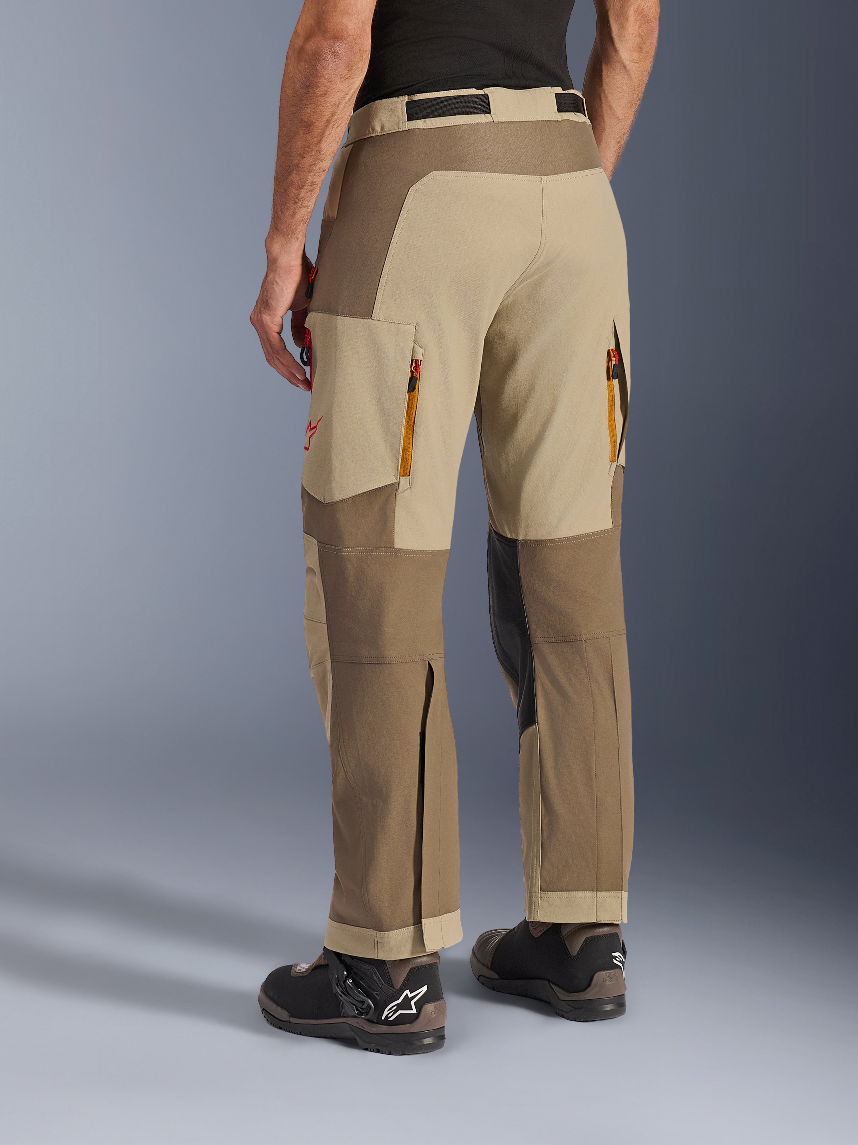 Pantaloni Alpinestars Maxdura Dual in Walnut Aluminum, vista laterale posteriore, pantaloni da moto adventure con pannelli sabbia e marroni, scarichi d'aria posteriori con cerniera, ampie tasche cargo e design sopra lo stivale, indossati con stivali neri e marroni