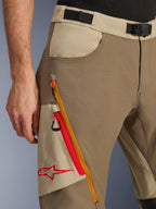 Alpinestars Maxdura Dual Pants in Walnut Aluminum, dettaglio ravvicinato che mostra la cintura in vita con fibbia magnetica, la tasca poggiamani e la tasca cargo angolata con cerniera con accenti rossi e arancioni e il logo Alpinestars.
