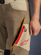 Alpinestars Maxdura Dual Pants, Walnut e Aluminum, primo piano del dettaglio sulla coscia con ampio sistema di ventilazione diagonale con cerniera e accenti rossi, logo Alpinestars rosso su pannello beige, abbigliamento moto adventure