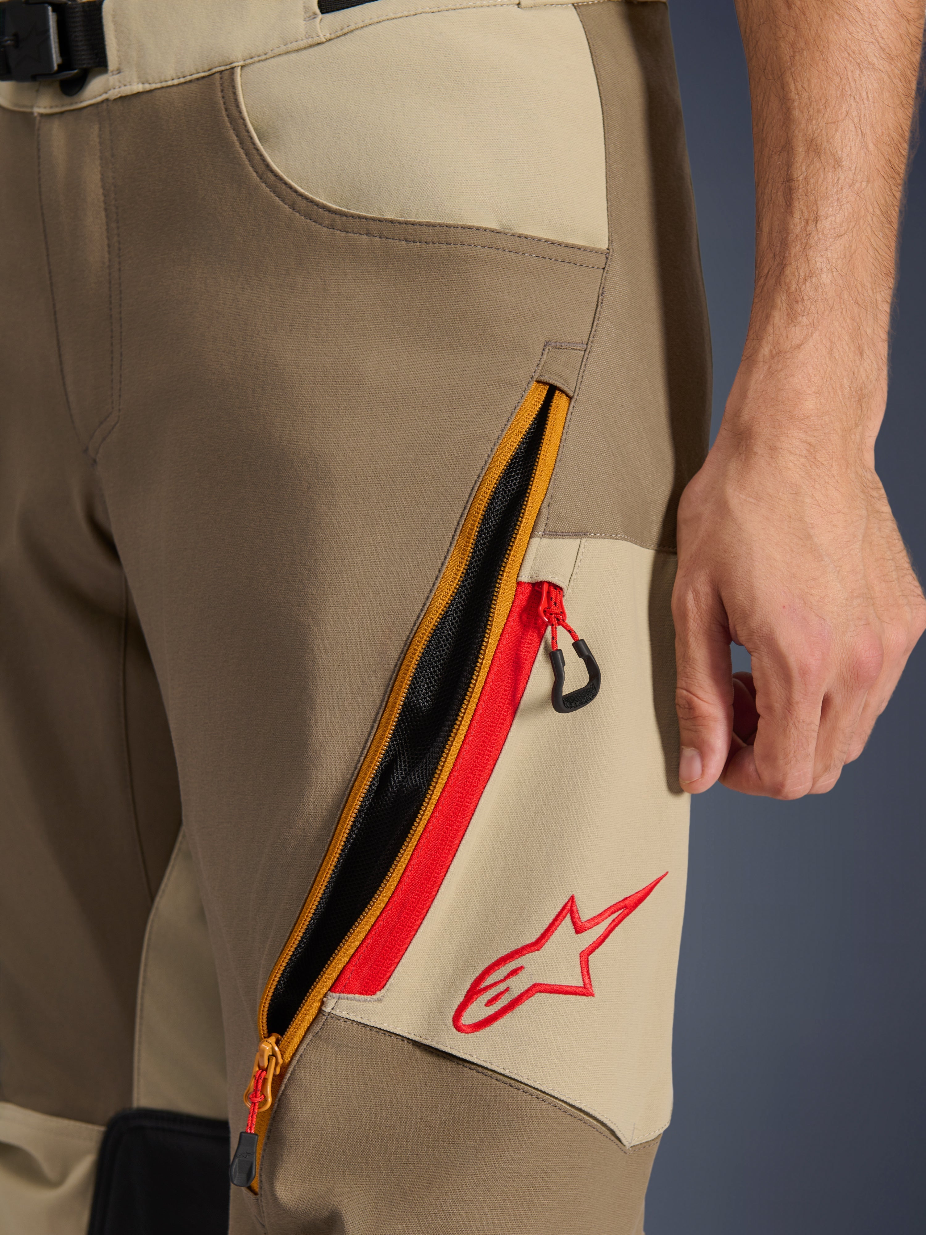 Alpinestars Maxdura Dual Pants, Walnut e Aluminum, primo piano del dettaglio sulla coscia con ampio sistema di ventilazione diagonale con cerniera e accenti rossi, logo Alpinestars rosso su pannello beige, abbigliamento moto adventure