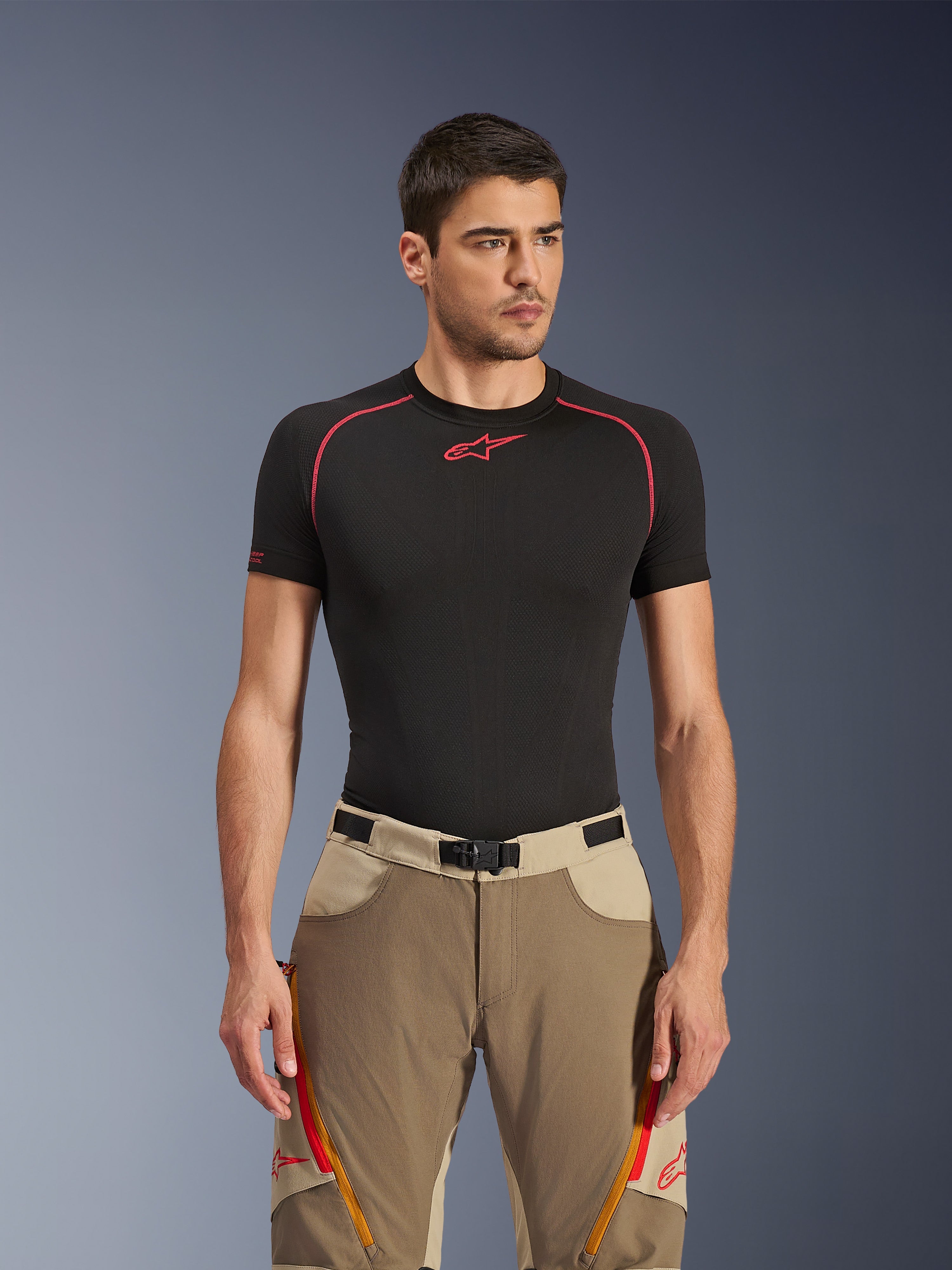 Alpinestars Maxdura Dual Pants in Walnut Aluminum, pantaloni da moto adventure marrone e beige, indossati da un modello con maglia tecnica nera, con prese d'aria sulle cosce con zip arancioni, loghi rossi e fibbia magnetica in vita, ritratto in studio.