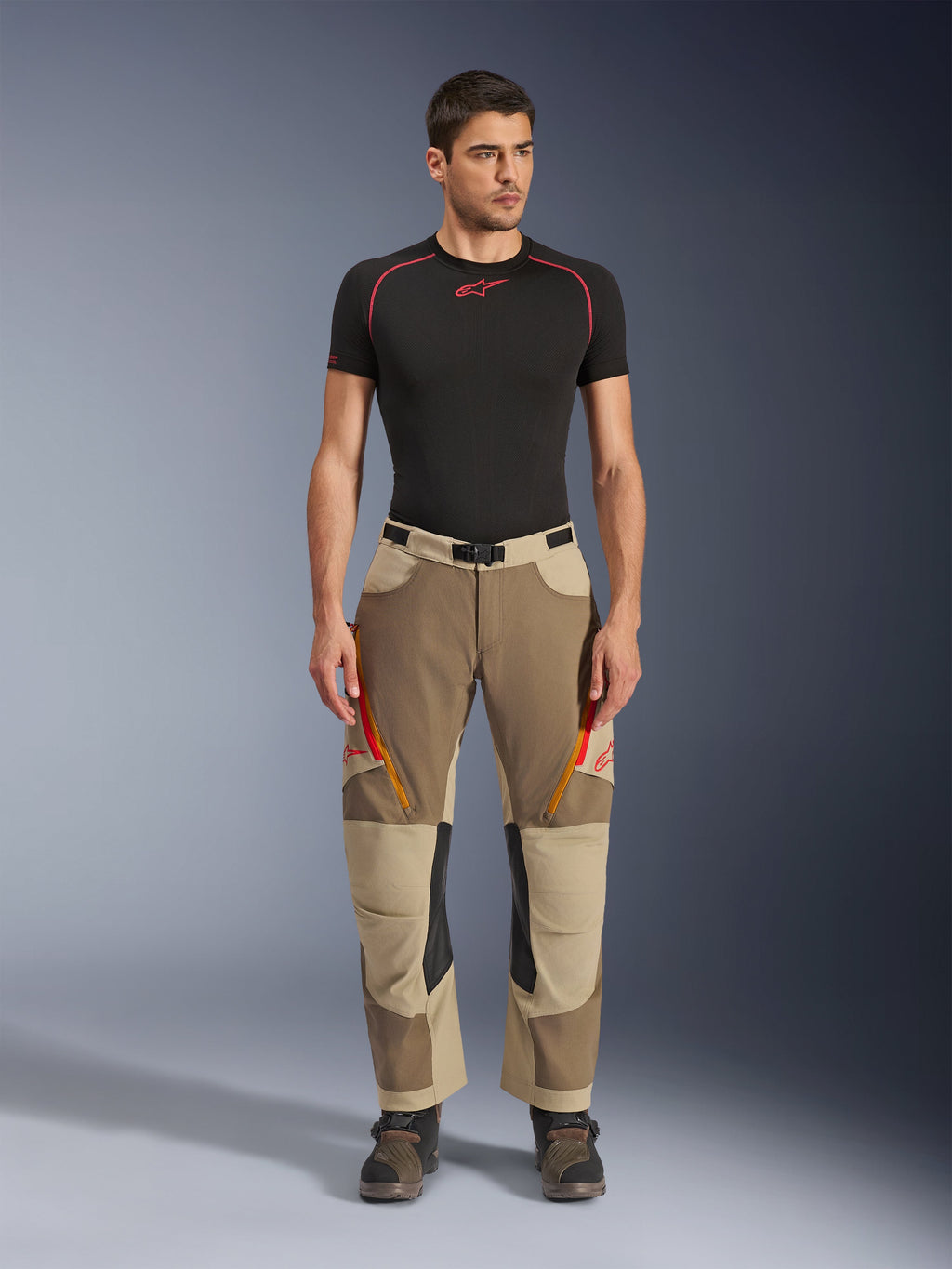 Alpinestars Maxdura Dual Pants, pantaloni da moto Adventure e Dual-Sport, Walnut Aluminum, indossati con una maglia a compressione nera a maniche corte e stivali adventure, caratterizzati da prese d'aria arancioni con cerniera e loghi rossi sulle cosce, cintura nera in vita con fibbia, design sopra lo stivale per la guida in moto.