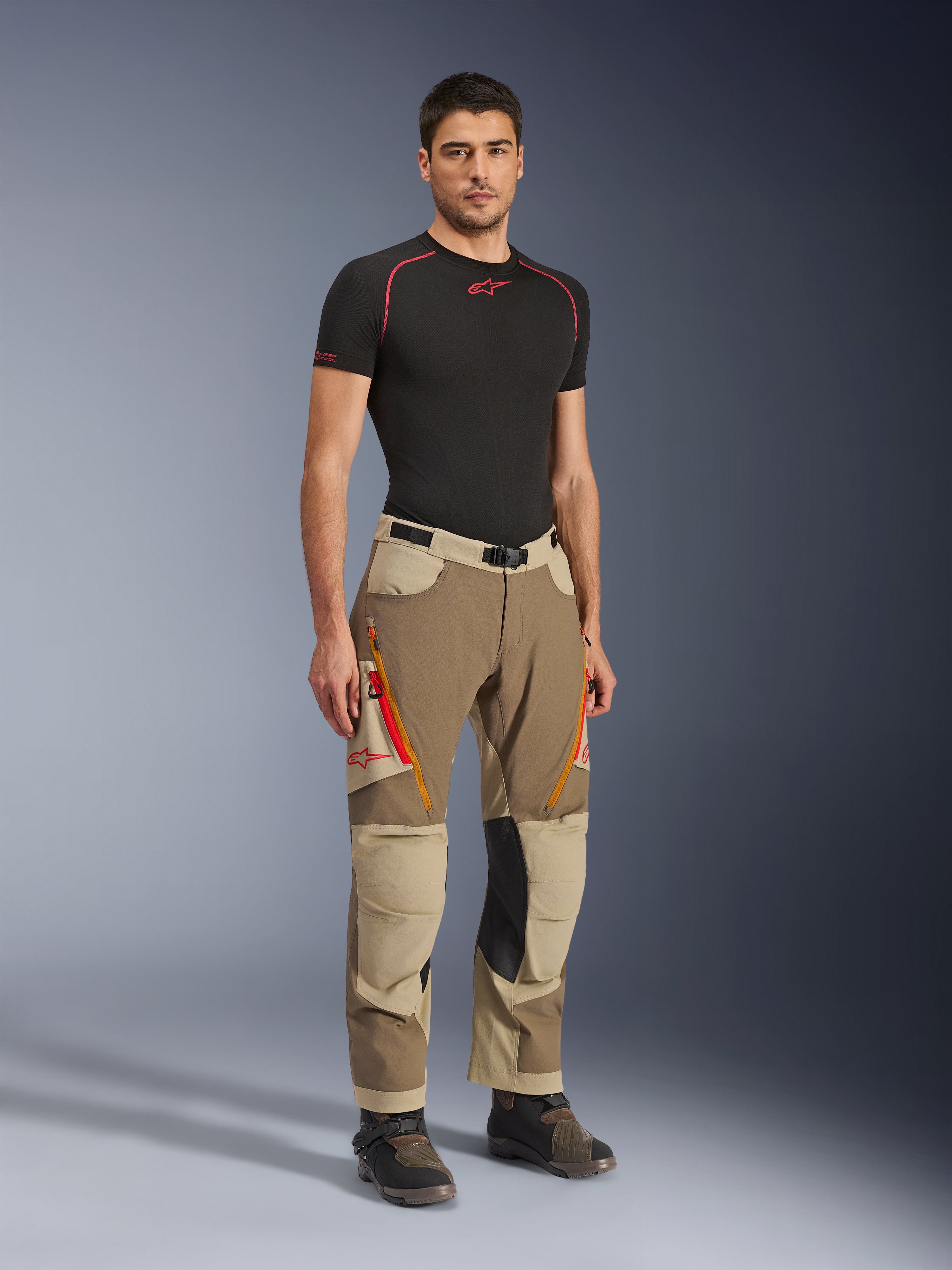 Alpinestars Maxdura Dual Pants in Walnut Aluminum, pantaloni da moto adventure marroni e beige con cerniere arancioni e inserti in pelle nera sulle ginocchia, indossati con t-shirt base layer nera e stivali adventure, vista frontale a figura intera