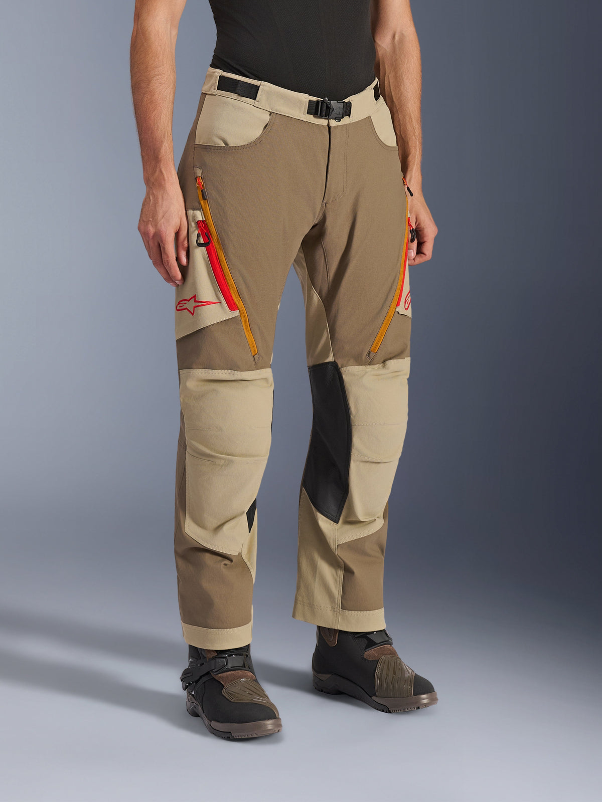 Pantaloni Alpinestars Maxdura Dual, pantaloni da moto adventure marrone e kaki, con prese d'aria sulle cosce con zip arancioni e rosse, pannelli interni alle ginocchia in pelle nera rinforzata e logo Astars rosso, indossati con stivali tecnici.