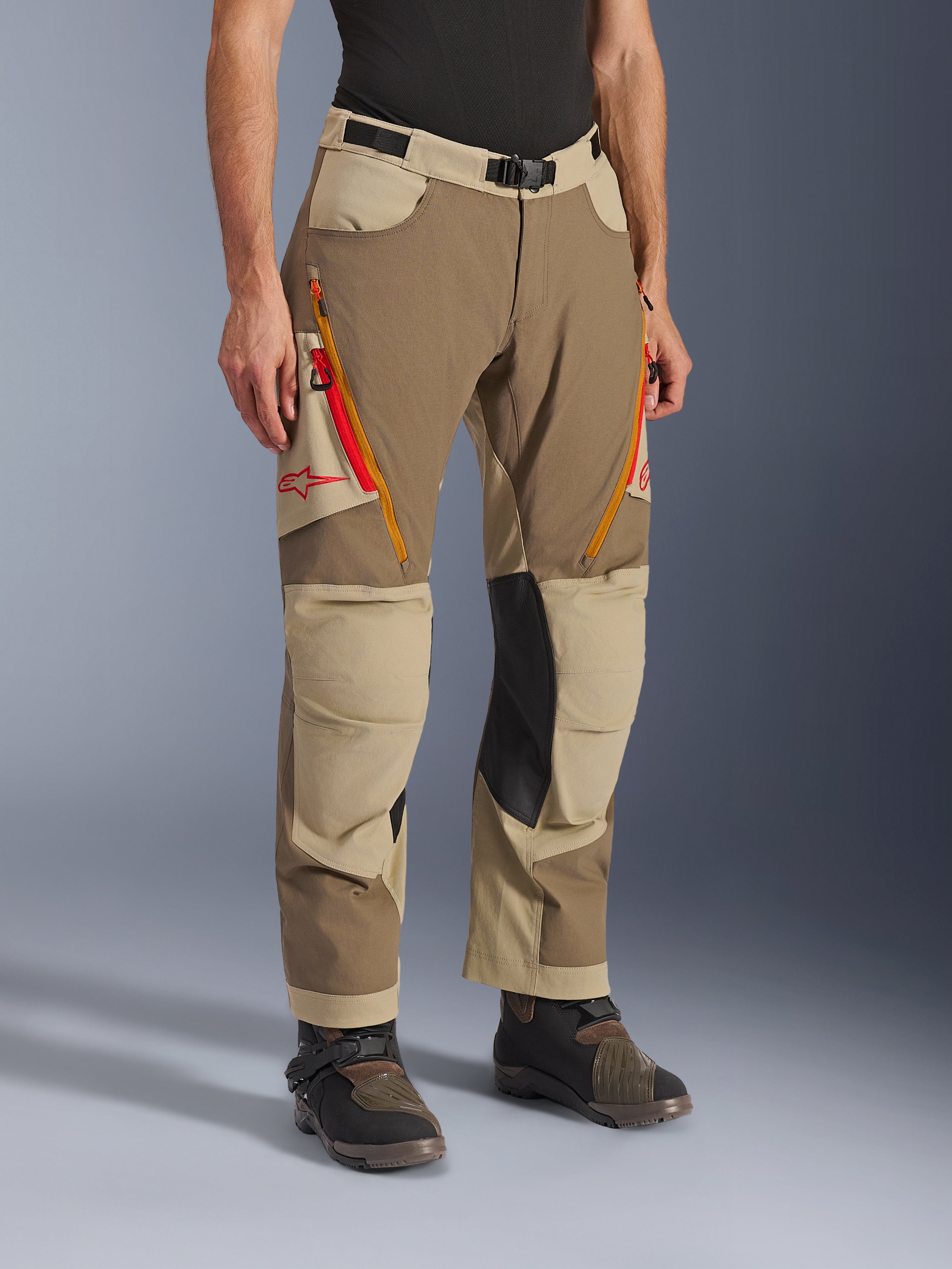 Pantaloni Alpinestars Maxdura Dual, pantaloni da moto adventure marrone e kaki, con prese d'aria sulle cosce con zip arancioni e rosse, pannelli interni alle ginocchia in pelle nera rinforzata e logo Astars rosso, indossati con stivali tecnici.