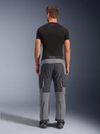 Alpinestars Maxdura Dual Pants, pantaloni da moto adventure, grigio scuro e grigio profondo, vista posteriore su modello con t-shirt nera e stivali, caratterizzati da pannelli grigi multitono e tasche cargo con zip e dettagli rossi.