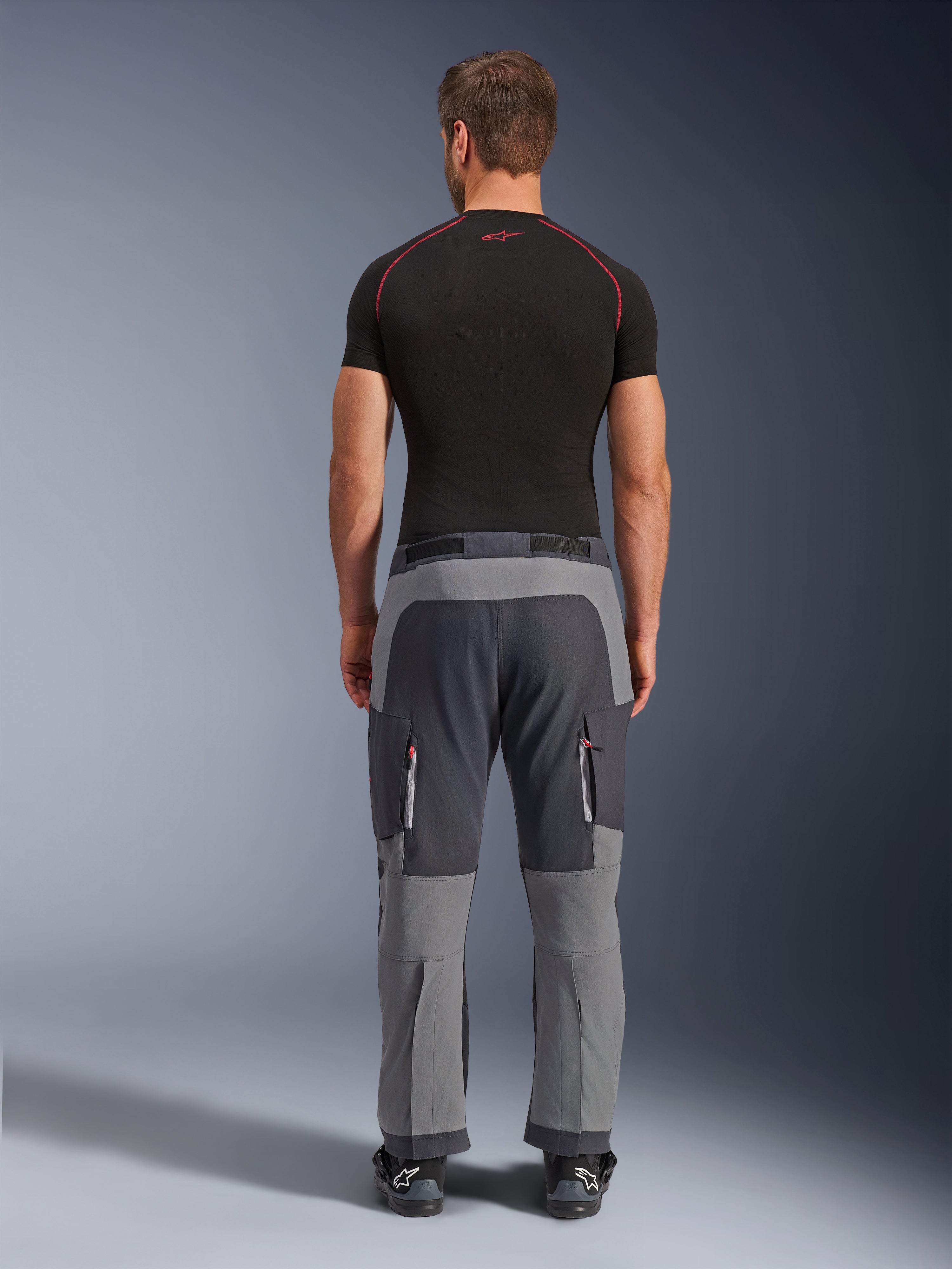 Alpinestars Maxdura Dual Pants, pantaloni da moto adventure, grigio scuro e grigio profondo, vista posteriore su modello con t-shirt nera e stivali, caratterizzati da pannelli grigi multitono e tasche cargo con zip e dettagli rossi.