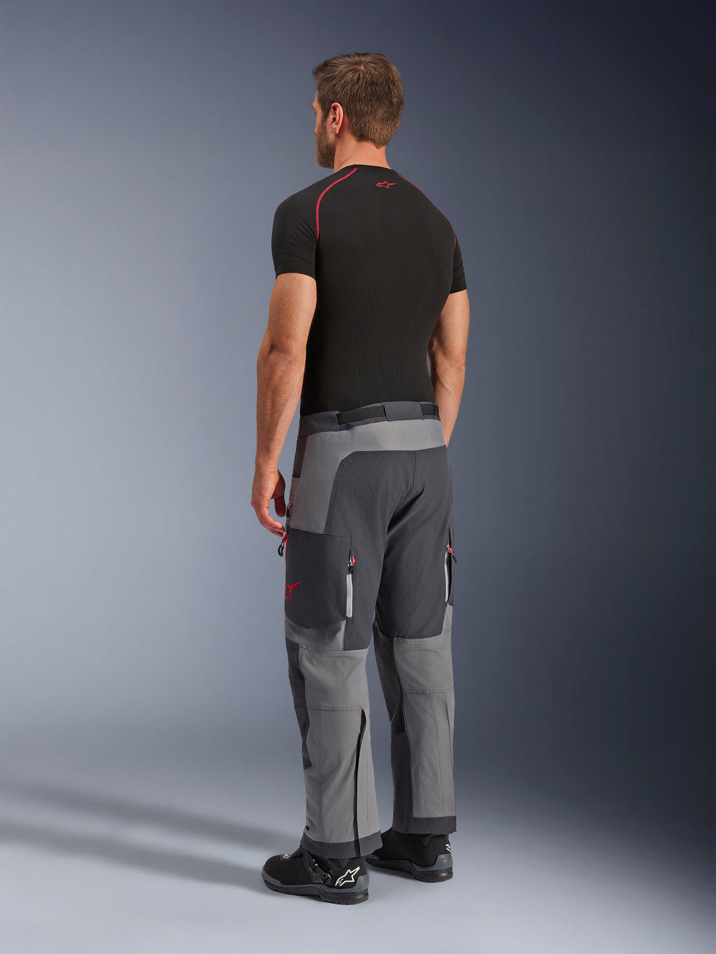 Alpinestars Maxdura Dual Pants, pantaloni da moto adventure, grigio scuro e grigio intenso, indossati da un modello in una vista posteriore laterale, con accenti rossi su cerniere e logo, pannelli grigi rinforzati, tasche cargo e vestibilità over-the-boot