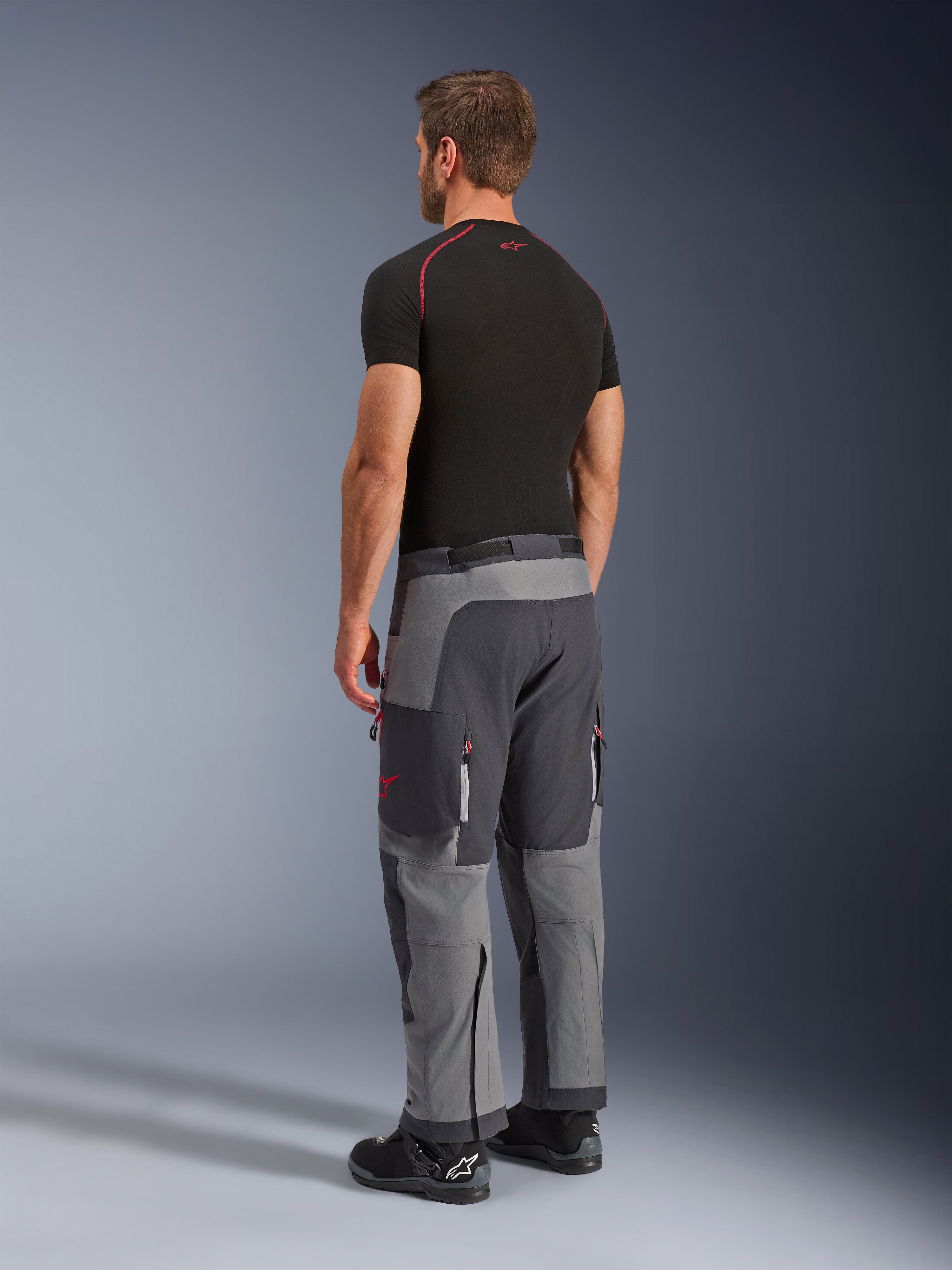 Alpinestars Maxdura Dual Pants, pantaloni da moto adventure, grigio scuro e grigio intenso, indossati da un modello in una vista posteriore laterale, con accenti rossi su cerniere e logo, pannelli grigi rinforzati, tasche cargo e vestibilità over-the-boot