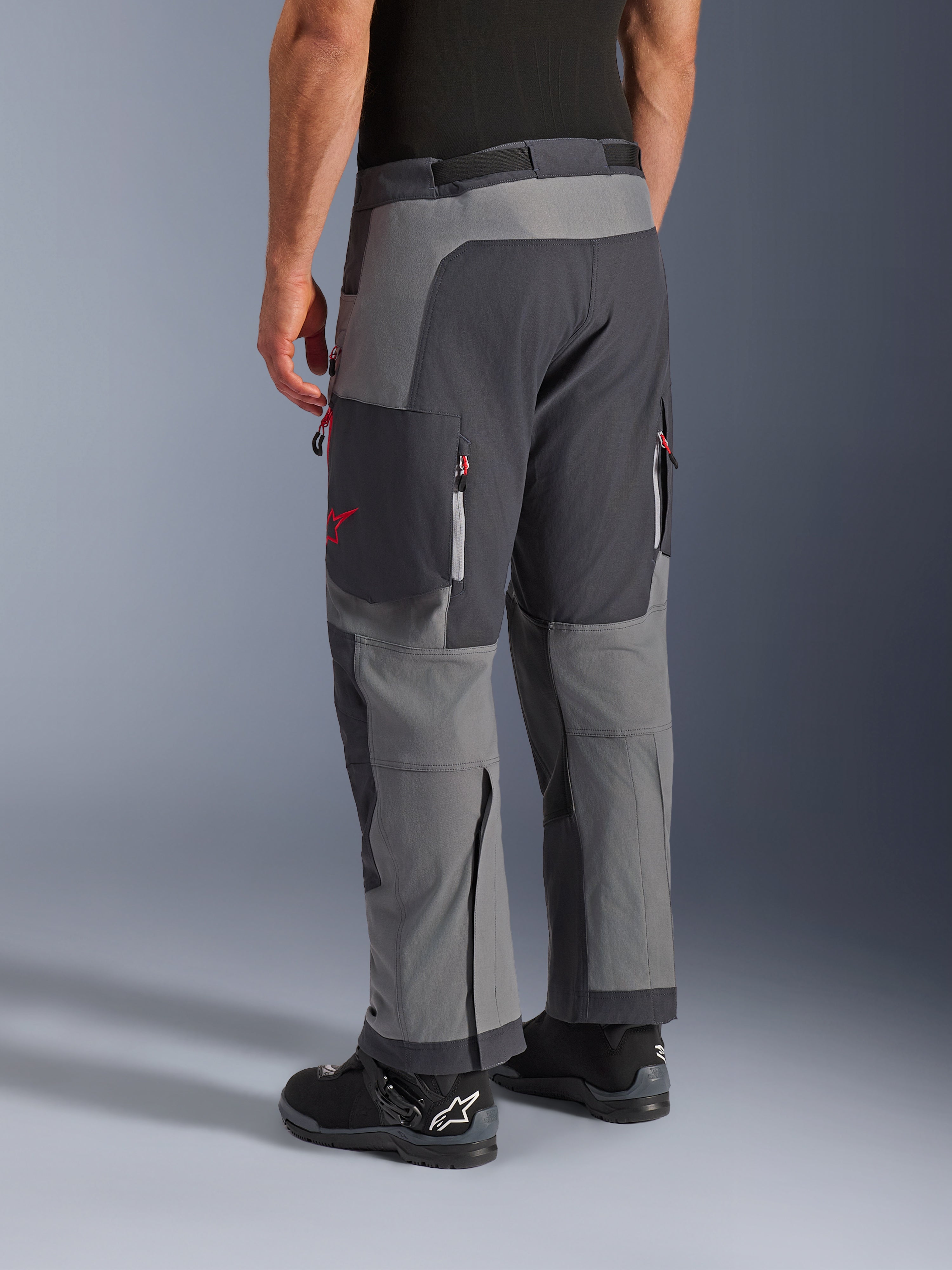 Pantaloni Alpinestars Maxdura Dual, pantaloni da moto adventure, grigio scuro e grigio intenso, vista posteriore con costruzione multi-pannello, dettagli con zip rosse e logo a stella, abbinati a stivali Alpinestars neri, progettati per prestazioni dual-sport