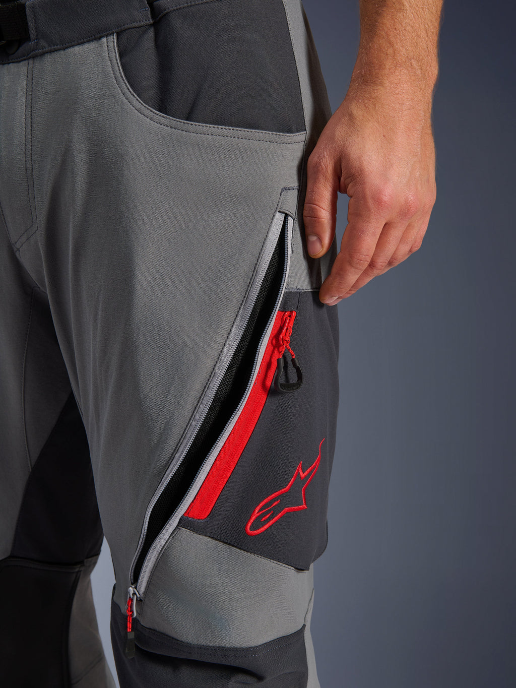 Pantaloni Alpinestars Maxdura Dual, pantaloni da moto adventure grigio scuro e grigio intenso, vista ravvicinata della presa d'aria sulla coscia con cerniera aperta, dettagli rossi e logo a stella rosso ricamato, design in tessuto tecnico elasticizzato