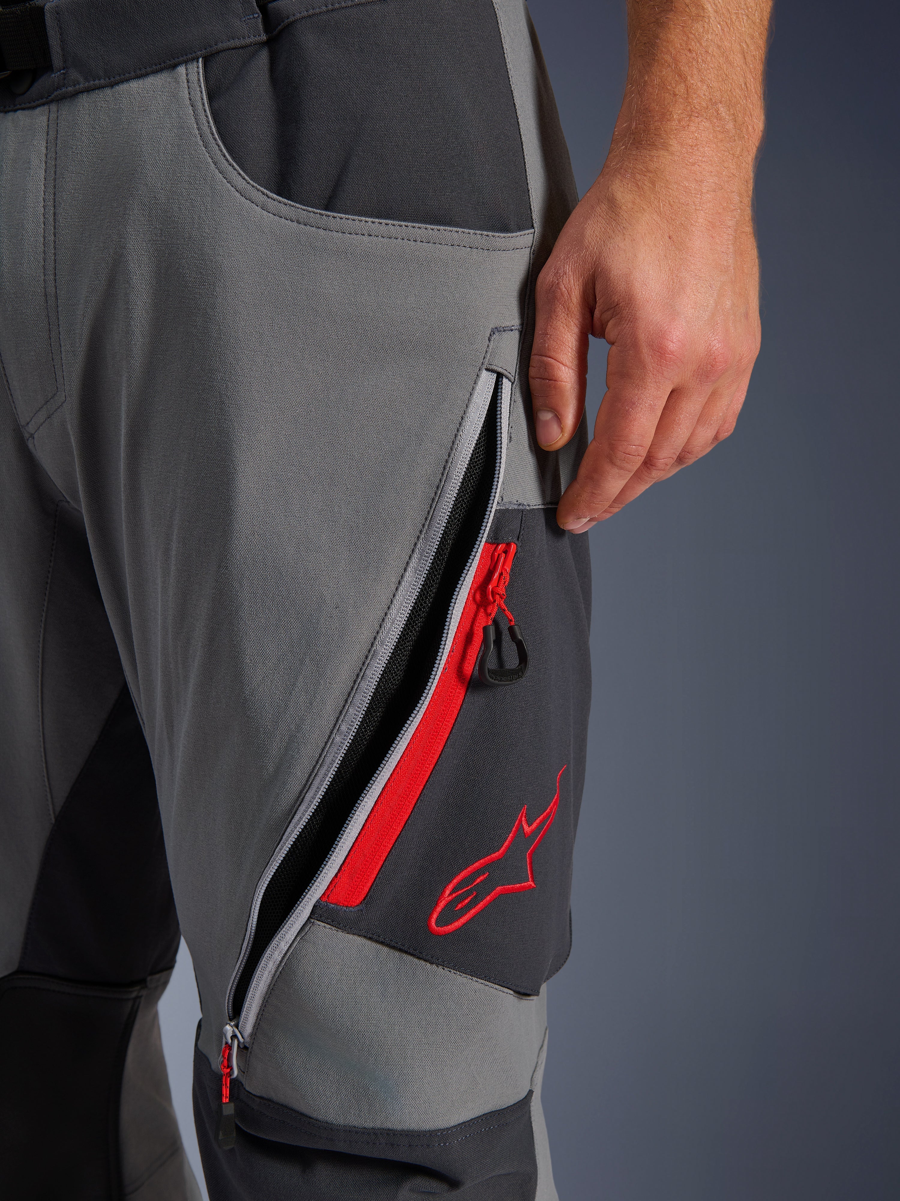 Pantaloni Alpinestars Maxdura Dual, pantaloni da moto adventure grigio scuro e grigio intenso, vista ravvicinata della presa d'aria sulla coscia con cerniera aperta, dettagli rossi e logo a stella rosso ricamato, design in tessuto tecnico elasticizzato