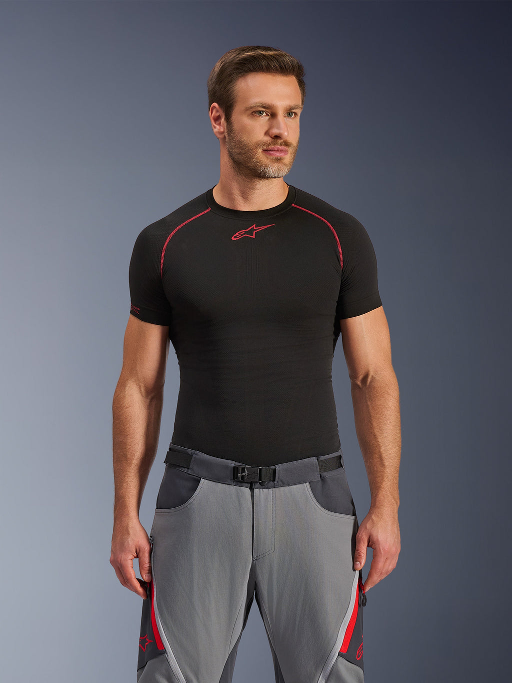 Pantaloni Alpinestars Maxdura Dual, grigio scuro e grigio intenso con accenti rossi, indossati con una maglia base layer nera con cuciture rosse, vestibilità atletica con cintura integrata a fibbia magnetica, abbigliamento resistente per moto adventure