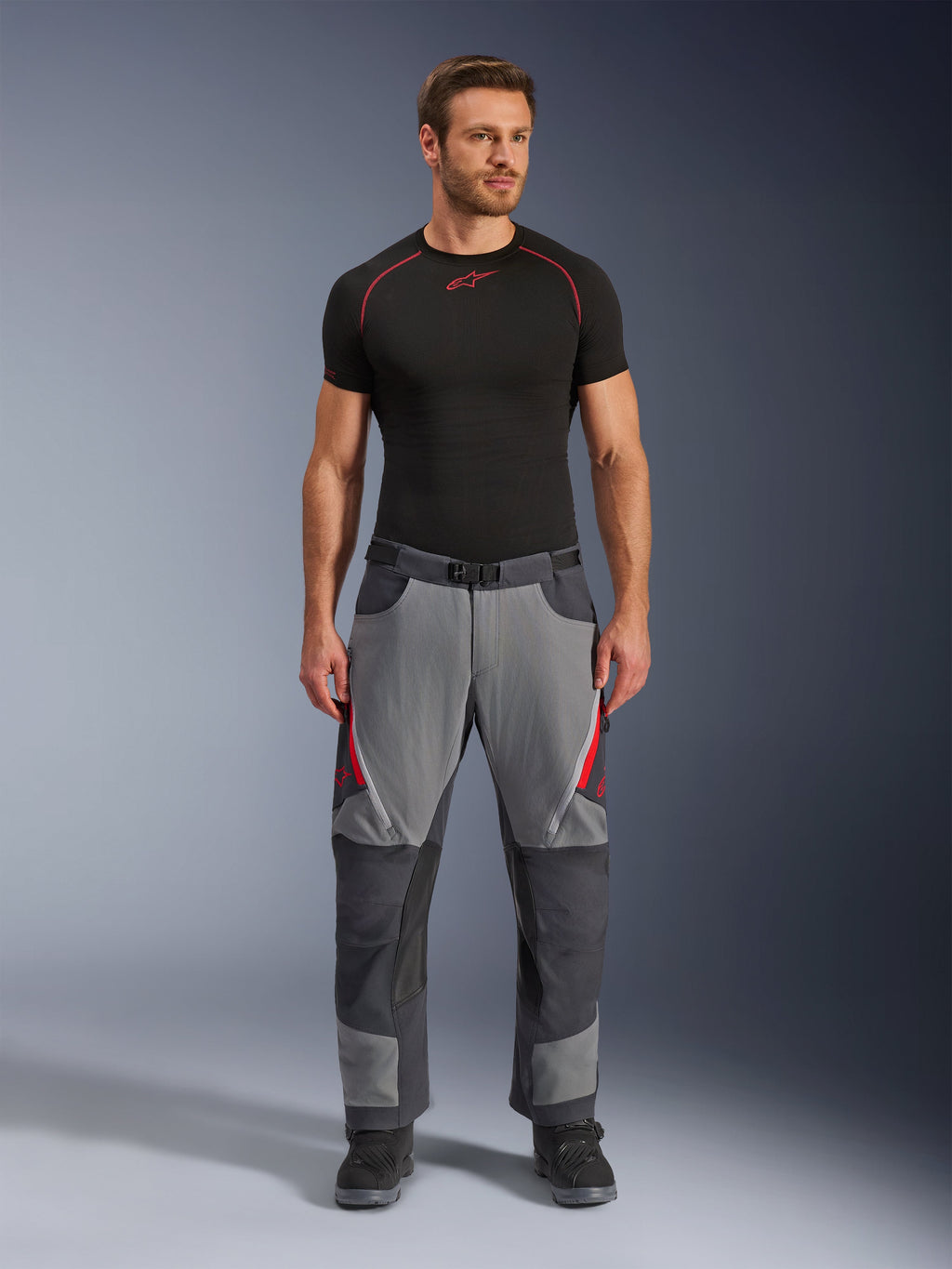 Alpinestars Maxdura Dual Pants, pantaloni da moto, grigio scuro e grigio intenso con accenti rossi, vista frontale indossati da un modello con base layer Alpinestars nero e stivali neri, caratterizzati da pannelli rinforzati e prese d'aria con cerniera sulle cosce, abbigliamento tecnico da adventure