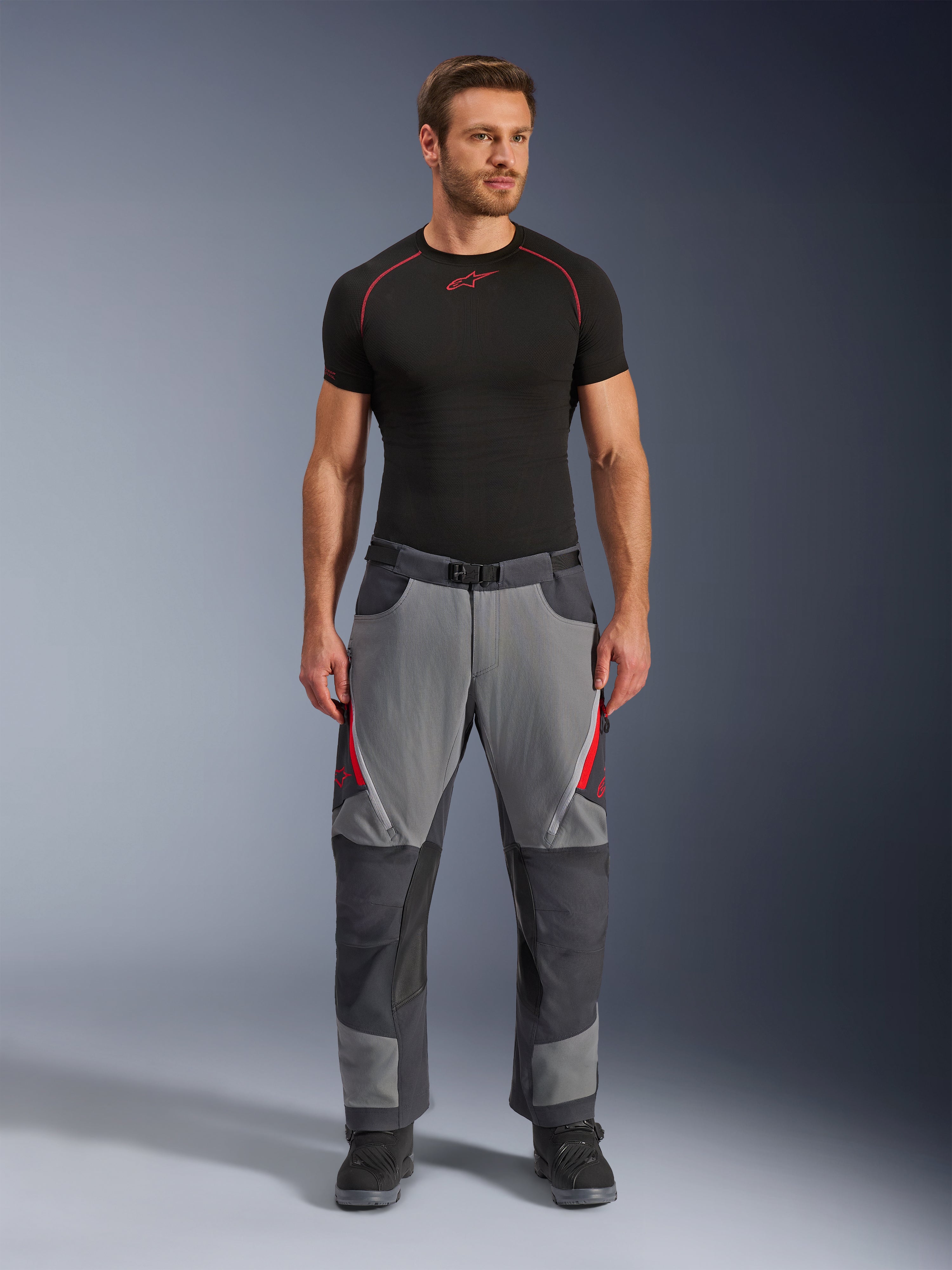 Alpinestars Maxdura Dual Pants, pantaloni da moto, grigio scuro e grigio intenso con accenti rossi, vista frontale indossati da un modello con base layer Alpinestars nero e stivali neri, caratterizzati da pannelli rinforzati e prese d'aria con cerniera sulle cosce, abbigliamento tecnico da adventure