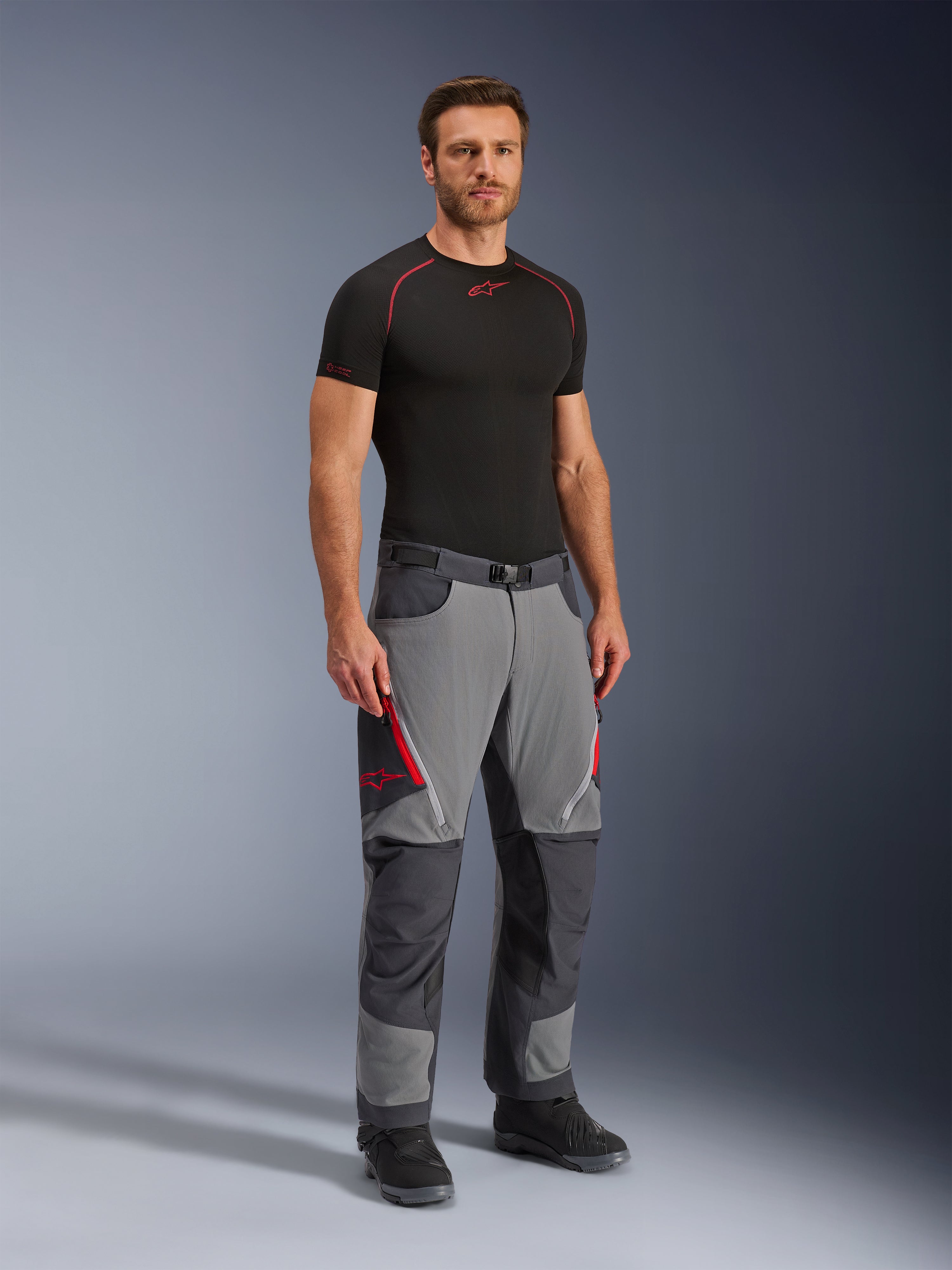 Alpinestars Maxdura Dual Pants, pantaloni da moto adventure, grigio scuro e grigio intenso, indossati con base layer nero e stivali, con prese d'aria rosse con zip, pannelli rinforzati su ginocchia e seduta, vita con cintura, vestibilità atletica per guida dual-sport