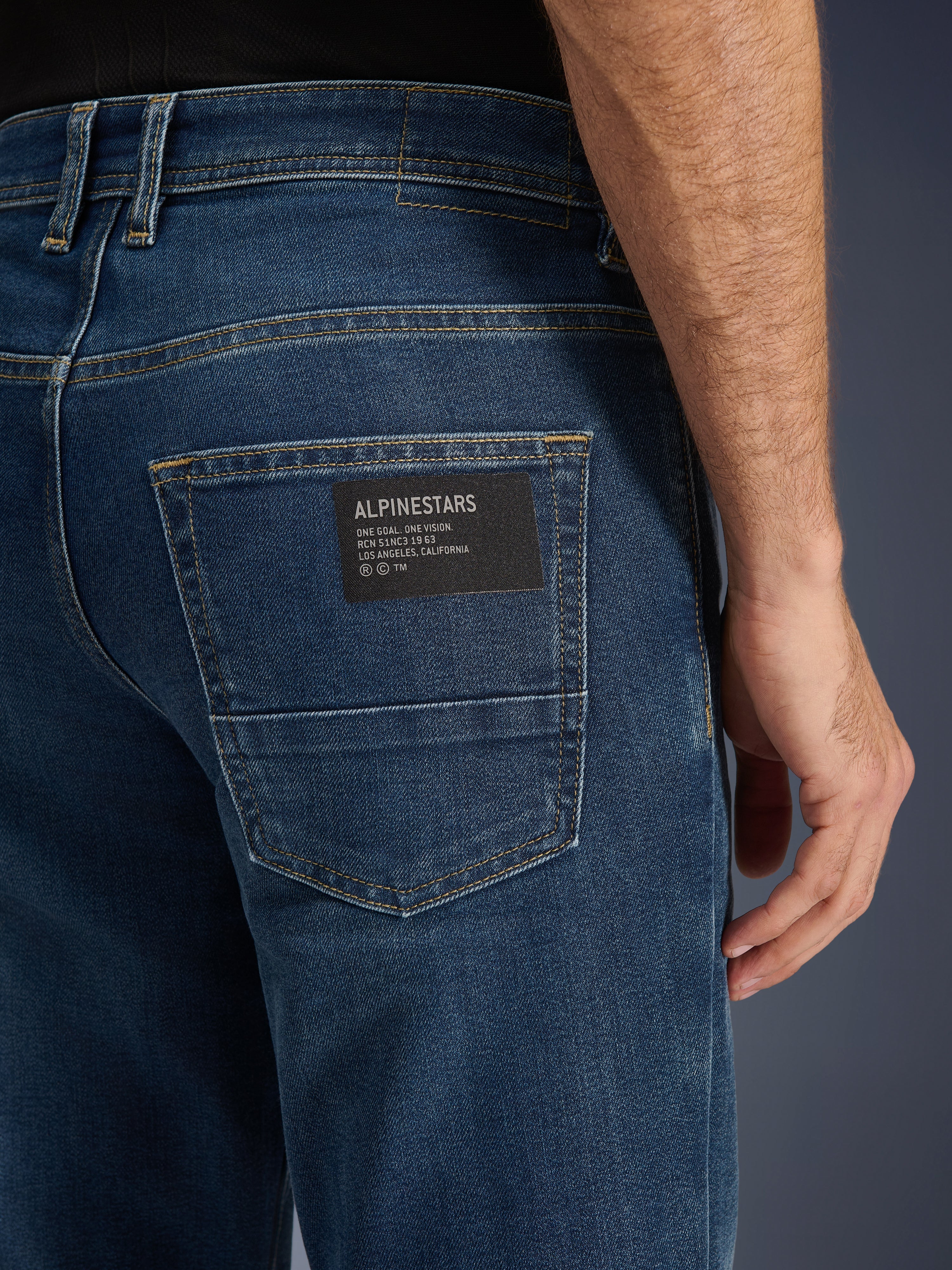 Alpinestars Flex-AST 5 Denim Pants, jeans da moto urban, lavaggio Indigo, primo piano della tasca posteriore con toppa nera del logo Alpinestars e testo 'One Goal. One Vision.', cuciture dorate a contrasto e trama del denim monostrato