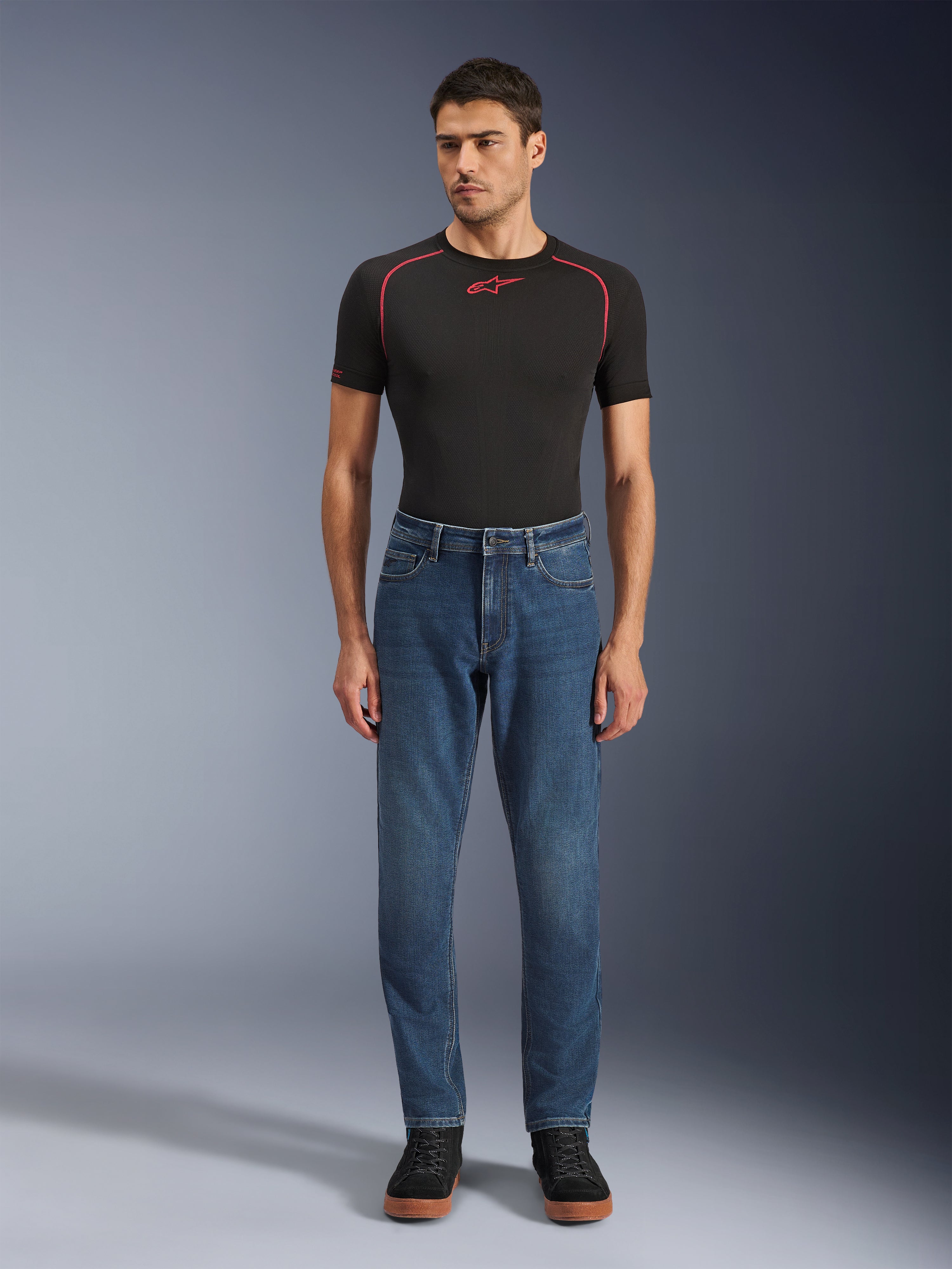 Pantaloni Alpinestars Flex-AST 5 Denim, pantaloni da moto in denim blu Indigo Washed, vestibilità affusolata regolare, indossati da un modello con un base layer Alpinestars nero e scarpe casual nere, abbigliamento urban riding lifestyle.