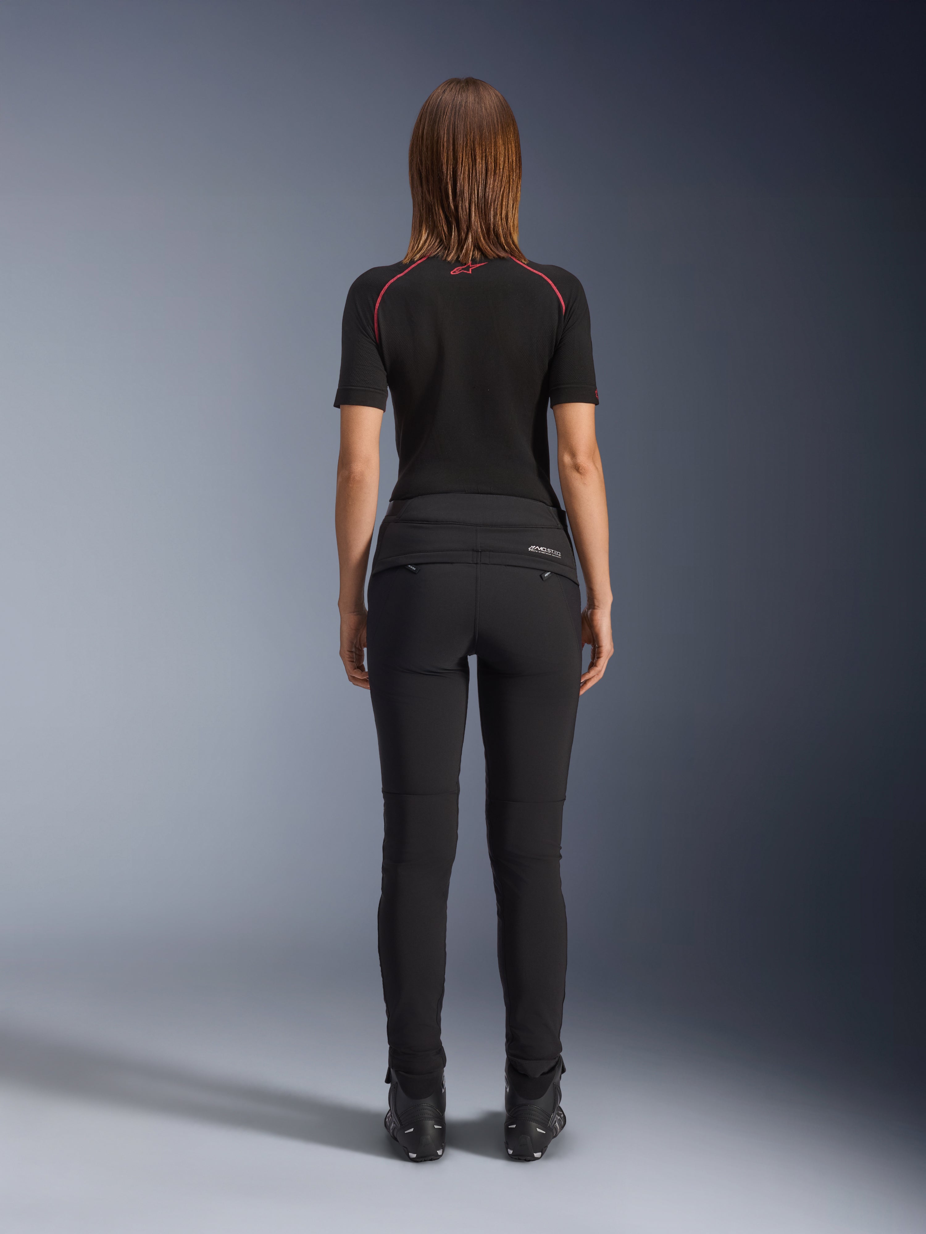 Alpinestars Alamosa Women Pants, nero, vista posteriore che mostra la struttura tessile slim-fit, tasche posteriori con cerniera e marchio MO.ST.EQ. riflettente sul carré posteriore, indossati con base layer nero a maniche corte e stivali da moto neri
