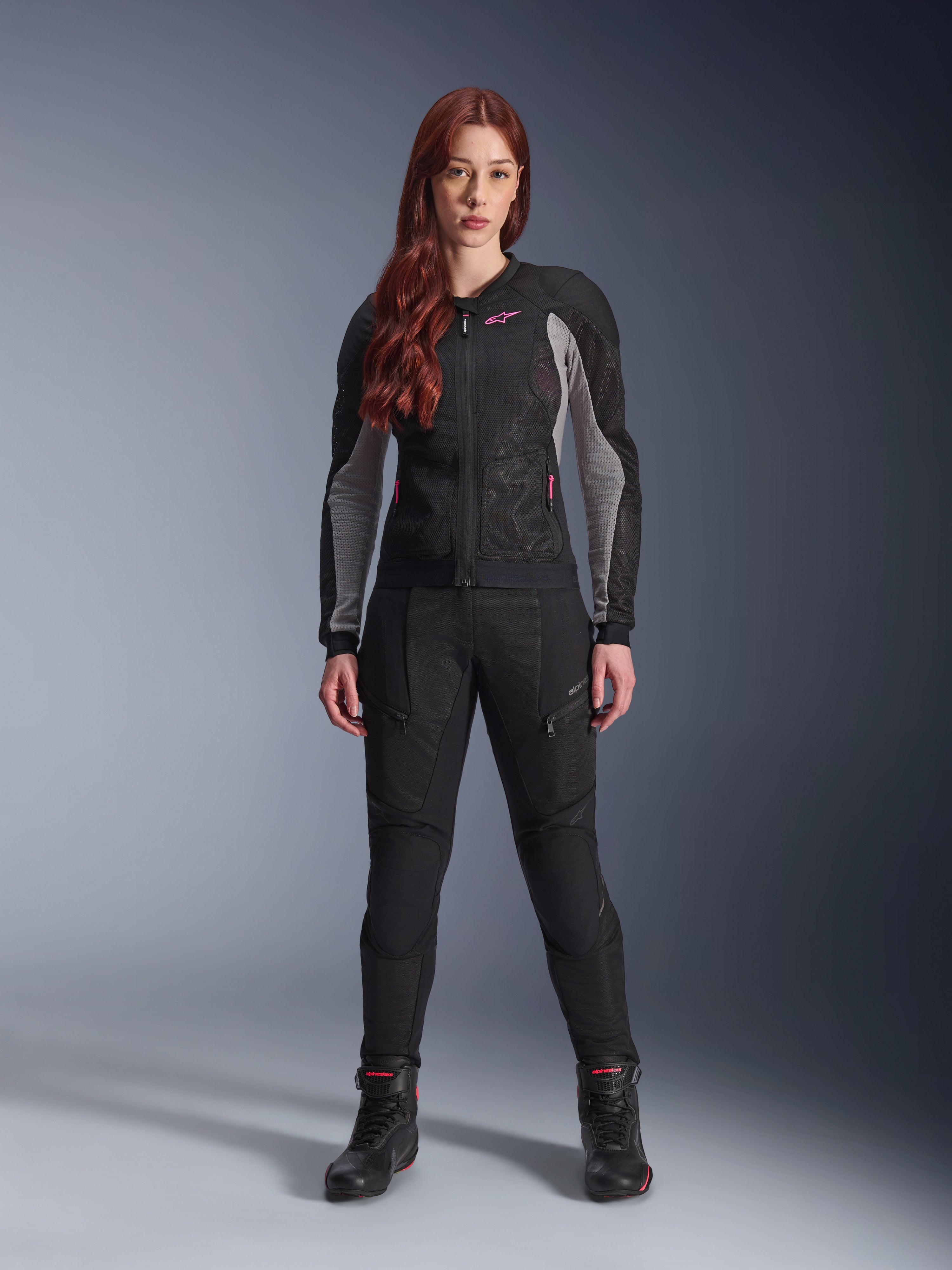 Giacca e pantaloni Alpinestars Stella Troop-Air, completo da moto per donna, nero con dettagli Ash Gray e Pink Fluo, indossati da una modella con capelli rossi, con ventilazione in mesh e design protettivo, posa in studio.