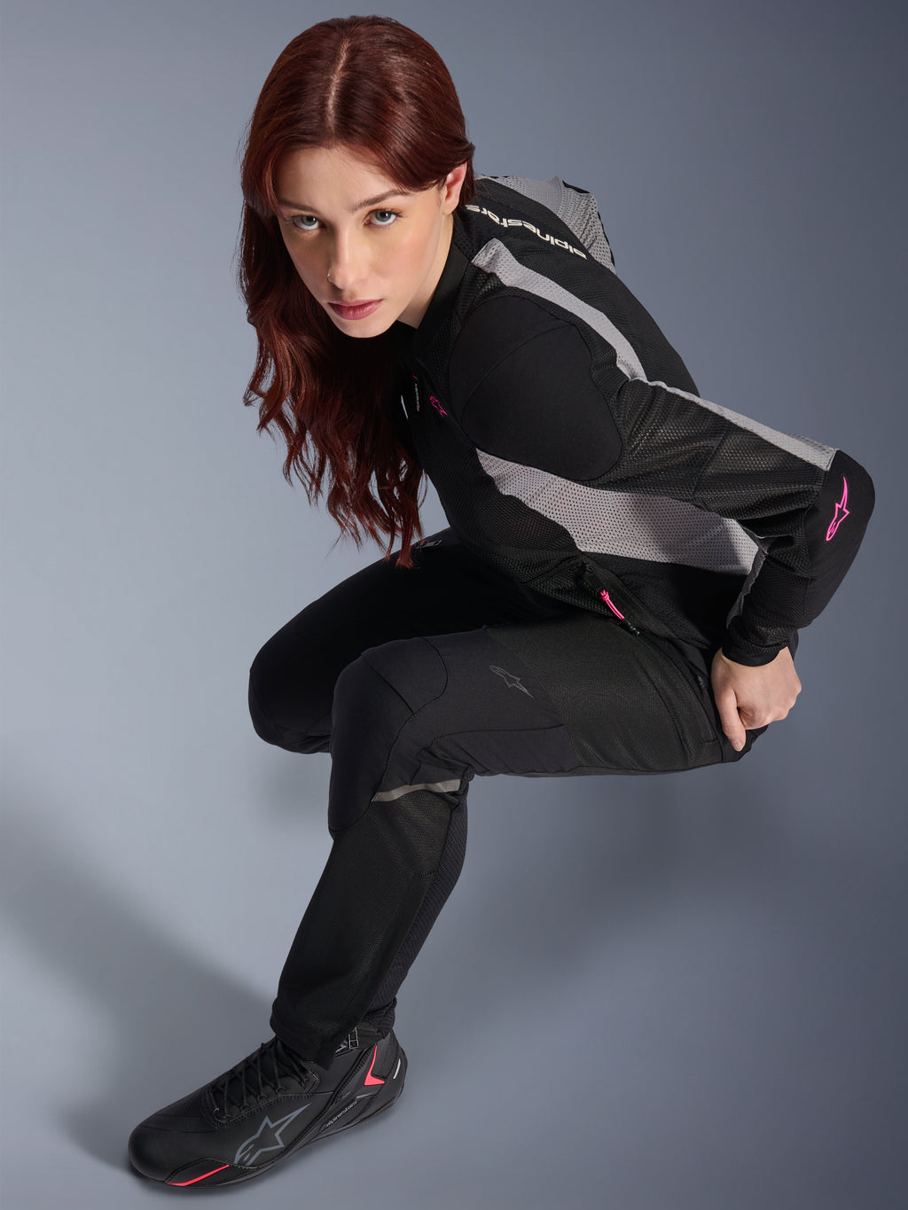 Donna che indossa i pantaloni Alpinestars Stella Troop-Air, neri, pantaloni in tessuto leggero e ventilato, abbinati a una giacca in mesh nera e grigia con dettagli rosa e stivali da moto neri, ritratto in studio, abbigliamento da moto urbano