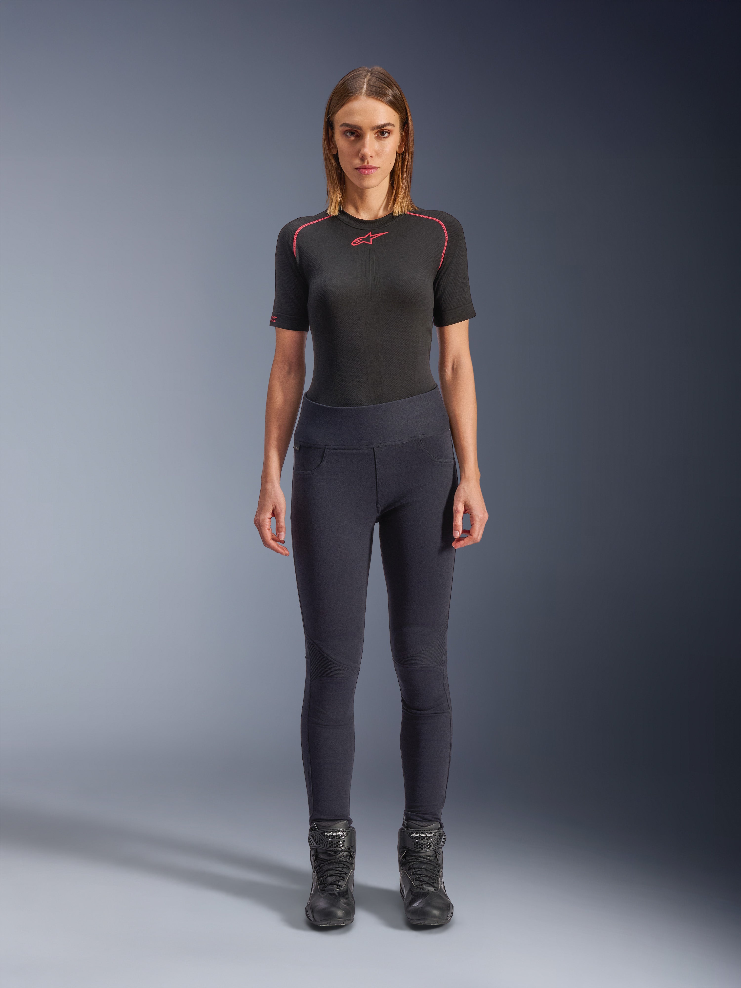 Leggings Alpinestars Stella Flex-AST, leggings da moto urban da donna in nero, design skinny a vita alta indossati con un top tecnico nero a maniche corte e stivali da moto neri, caratterizzati da pannelli a fisarmonica sulle ginocchia e un look minimalista contemporaneo