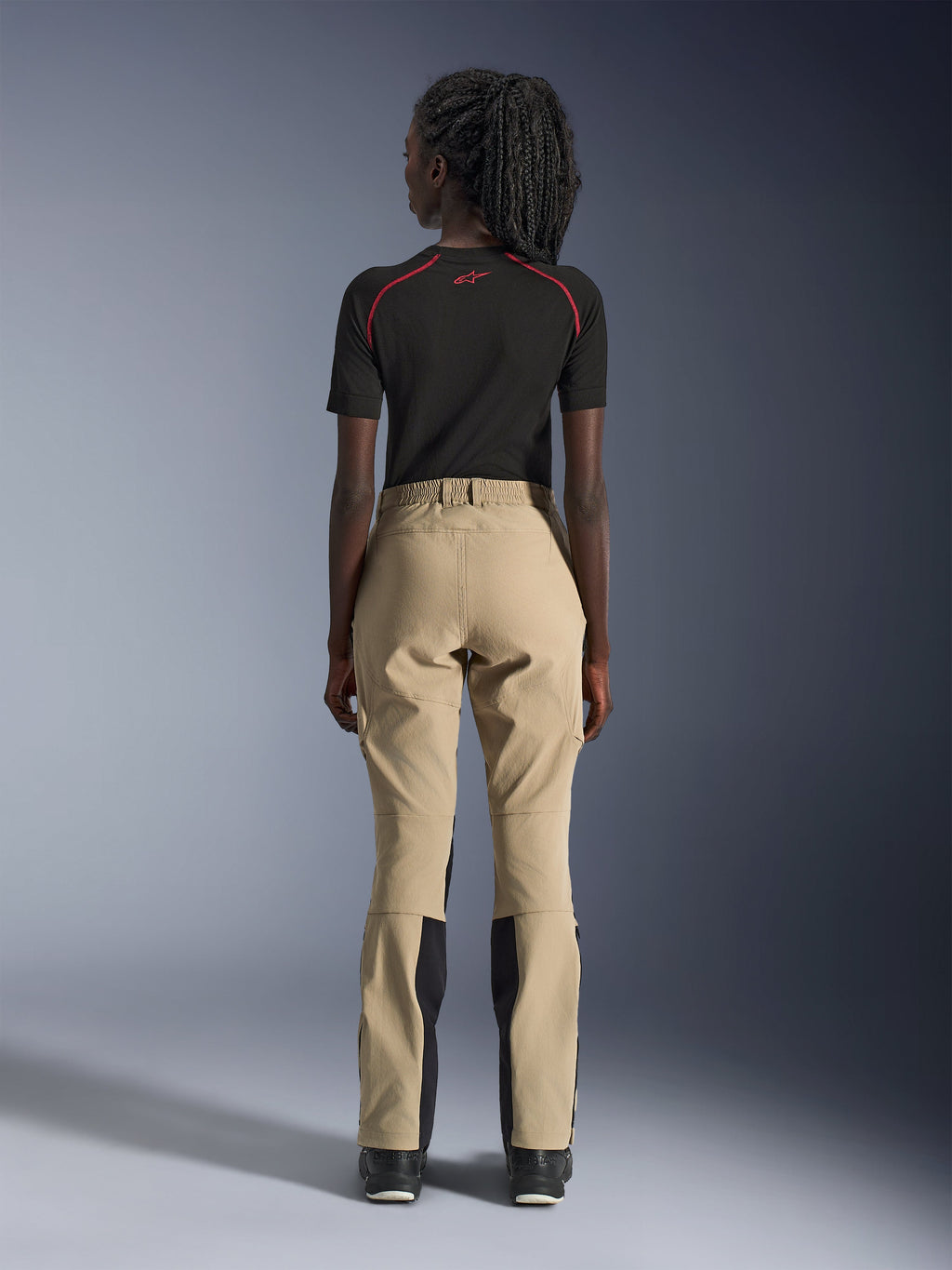 Pantaloni Alpinestars Woman Stella Flex-AST Explorer in Sand, vista posteriore su modella, abbinati a un base layer nero a maniche corte e stivali neri, con pannelli rinforzati sulla seduta e costruzione ergonomica in tessuto per adventure touring