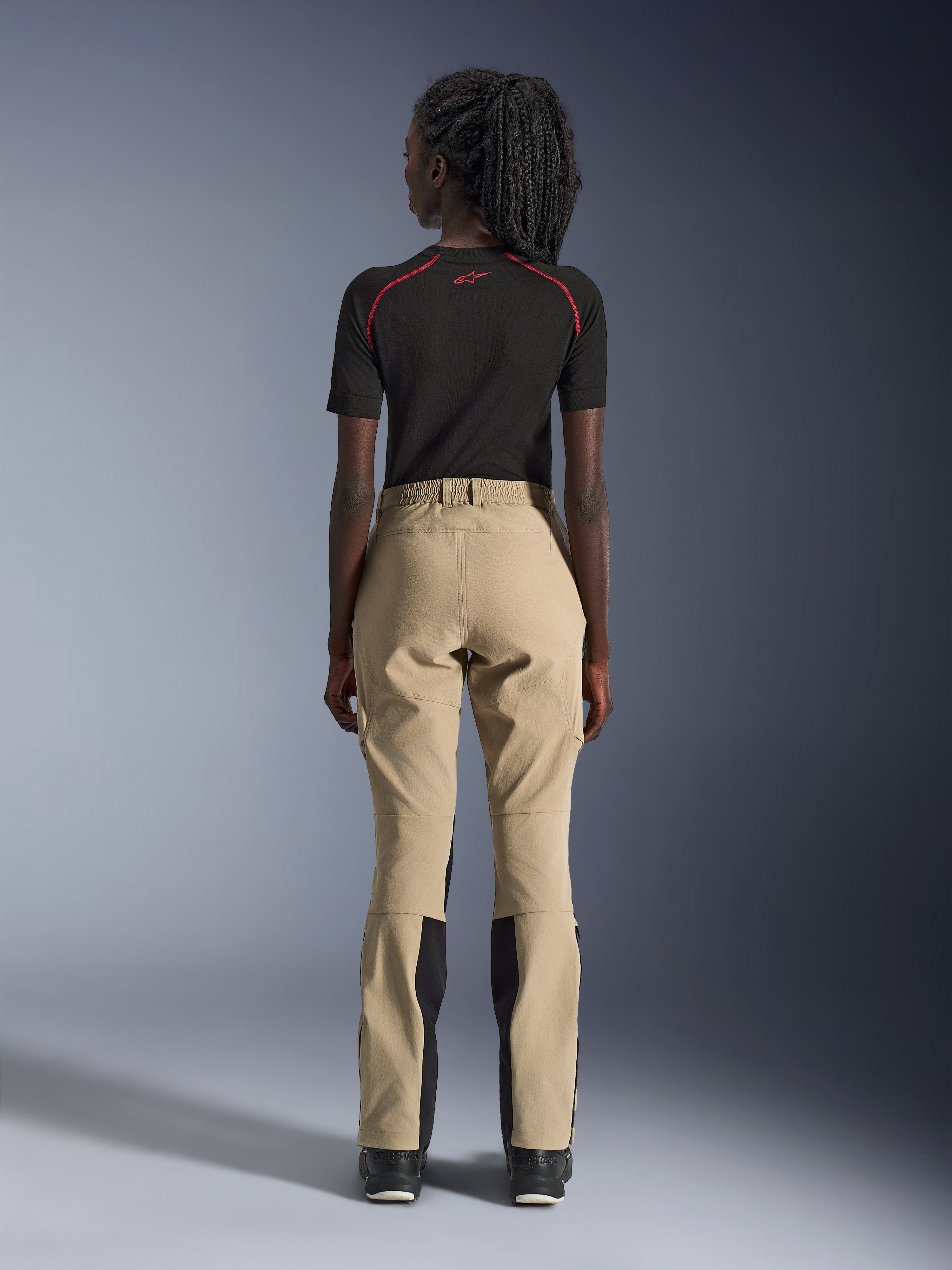 Pantaloni Alpinestars Woman Stella Flex-AST Explorer in Sand, vista posteriore su modella, abbinati a un base layer nero a maniche corte e stivali neri, con pannelli rinforzati sulla seduta e costruzione ergonomica in tessuto per adventure touring