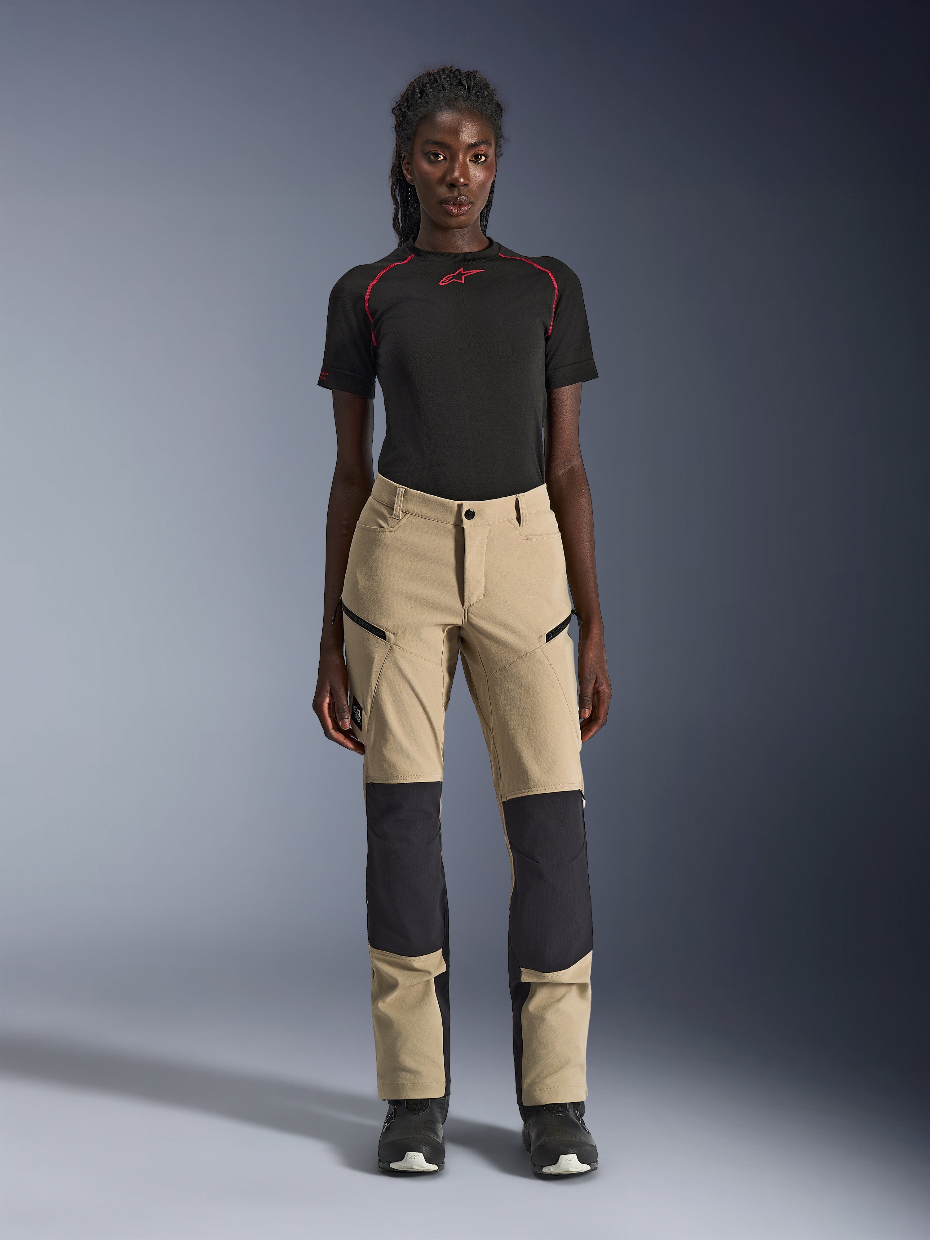 Pantaloni Alpinestars Stella Flex-AST Explorer da donna, pantaloni da moto, color sabbia con pannelli neri, indossati da una modella con maglia nera a maniche corte e stivali neri, dotati di pannelli rinforzati sulle ginocchia, tasche cargo con cerniera e tessuto ripstop traspirante per la guida adventure in climi caldi