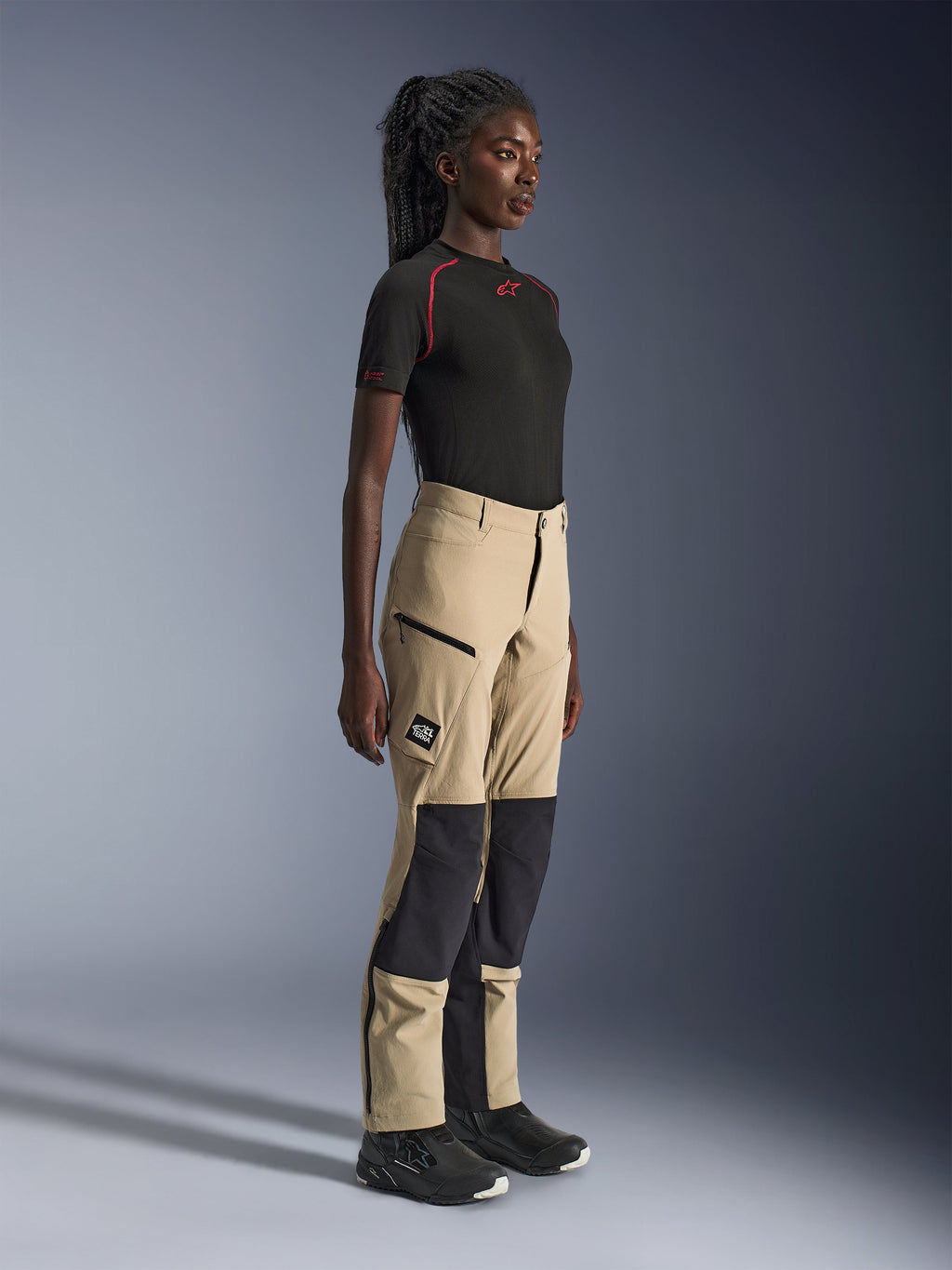 Pantaloni Alpinestars Stella Flex-AST Explorer da donna, pantaloni adventure touring sabbia e nero, profilo laterale su una modella che indossa una t-shirt Alpinestars nera e stivali da moto, dotati di rinforzi in ripstop sulle ginocchia e tasche cargo con cerniera.