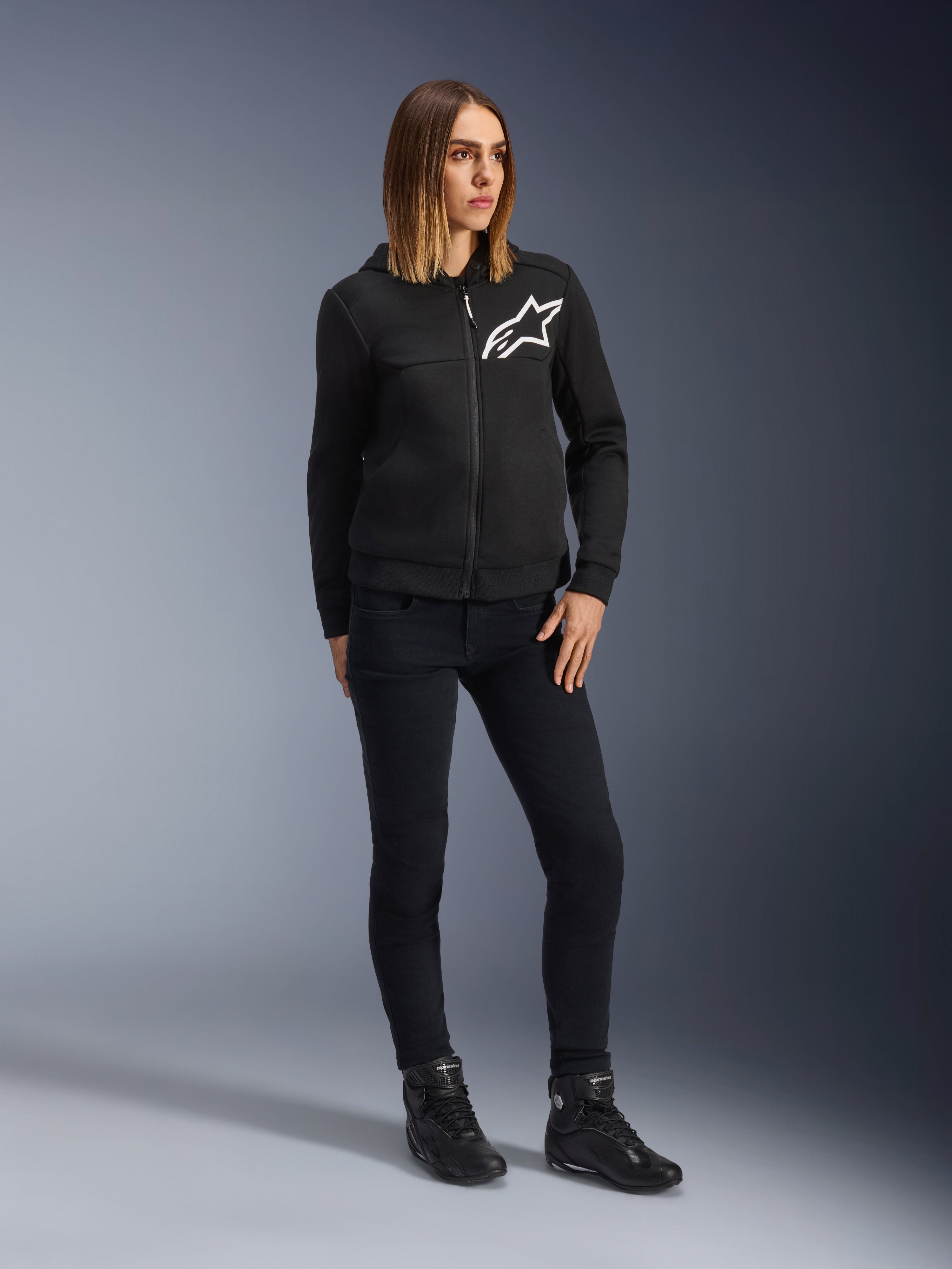Donna che indossa Alpinestars Daisy V3 Women's Riding Denim, pantaloni da moto slim-fit in Rinse Black, abbinati a una felpa nera con zip e stivali da moto, abbigliamento protettivo casual urbano