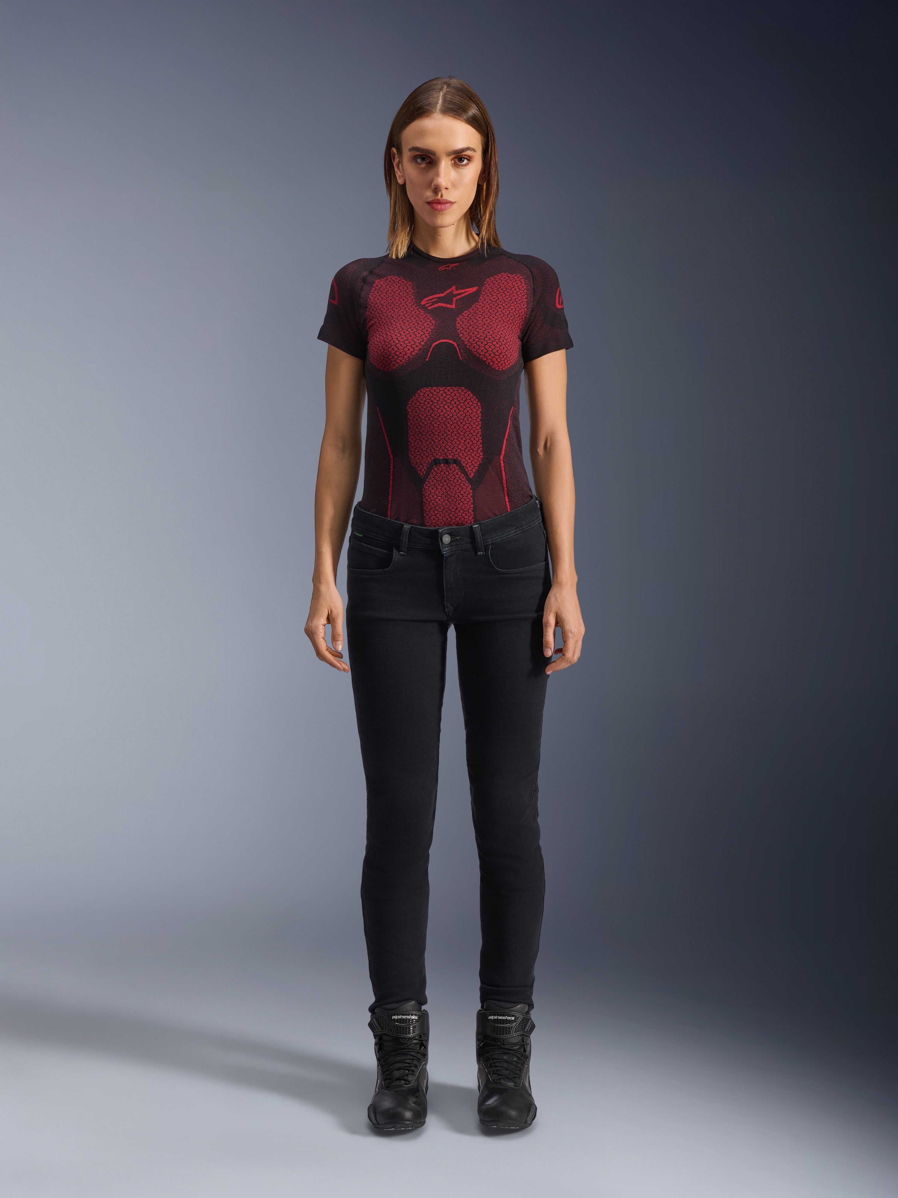 Alpinestars Daisy V3 Women's Riding Denim, pantaloni da moto slim-fit, nero, indossati da una modella con una t-shirt tecnica a motivi rossi e neri e stivali da moto neri, abbigliamento urban riding casual con silhouette femminile e design a vita alta.