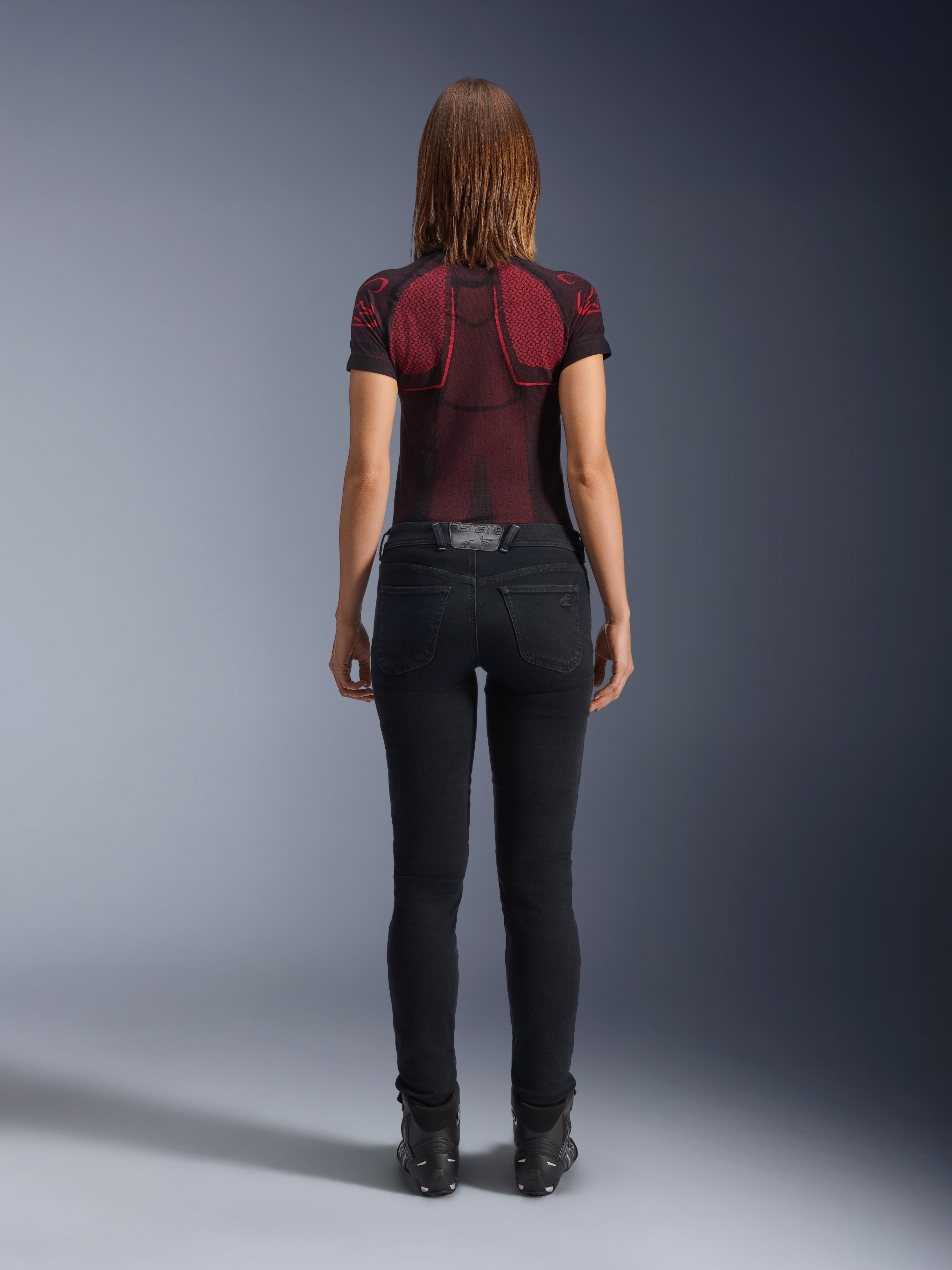 Alpinestars Daisy V3 Women's Riding Denim, Rinse Black, pantaloni da moto slim fit visti da dietro, con design a vita alta e tasche posteriori, abbinati a maglia tecnica rossa e nera e stivali neri, abbigliamento moto urban da donna
