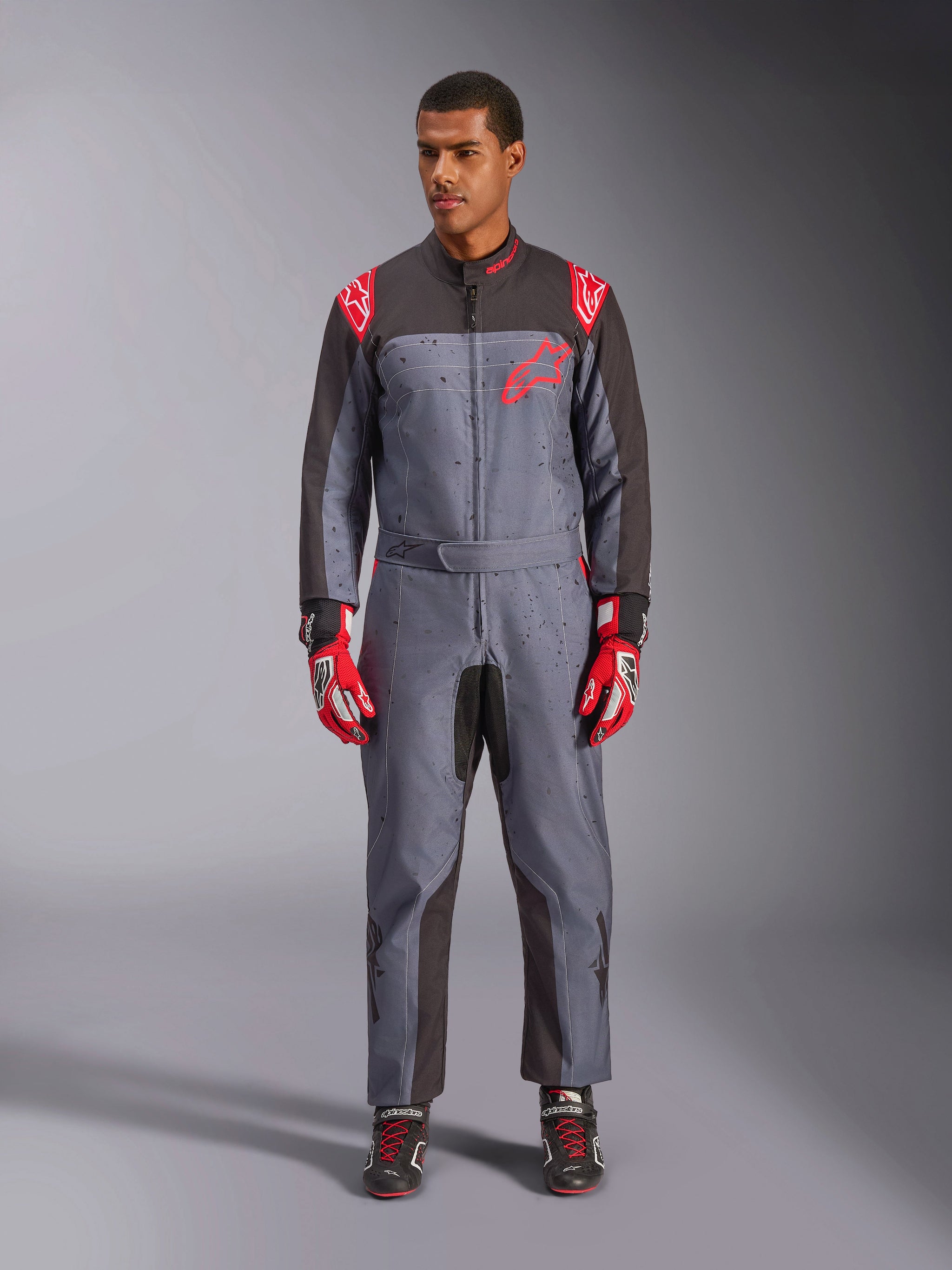 Tuta Alpinestars KMX-9 V3 Graphic 6, tuta da kart, antracite, nero e rosso, motivo grafico grigio maculato, pannelli neri sull'interno di braccia e gambe, loghi Alpinestars rossi sulle spalle e sul petto, indossata con guanti da corsa rossi e scarpe nere