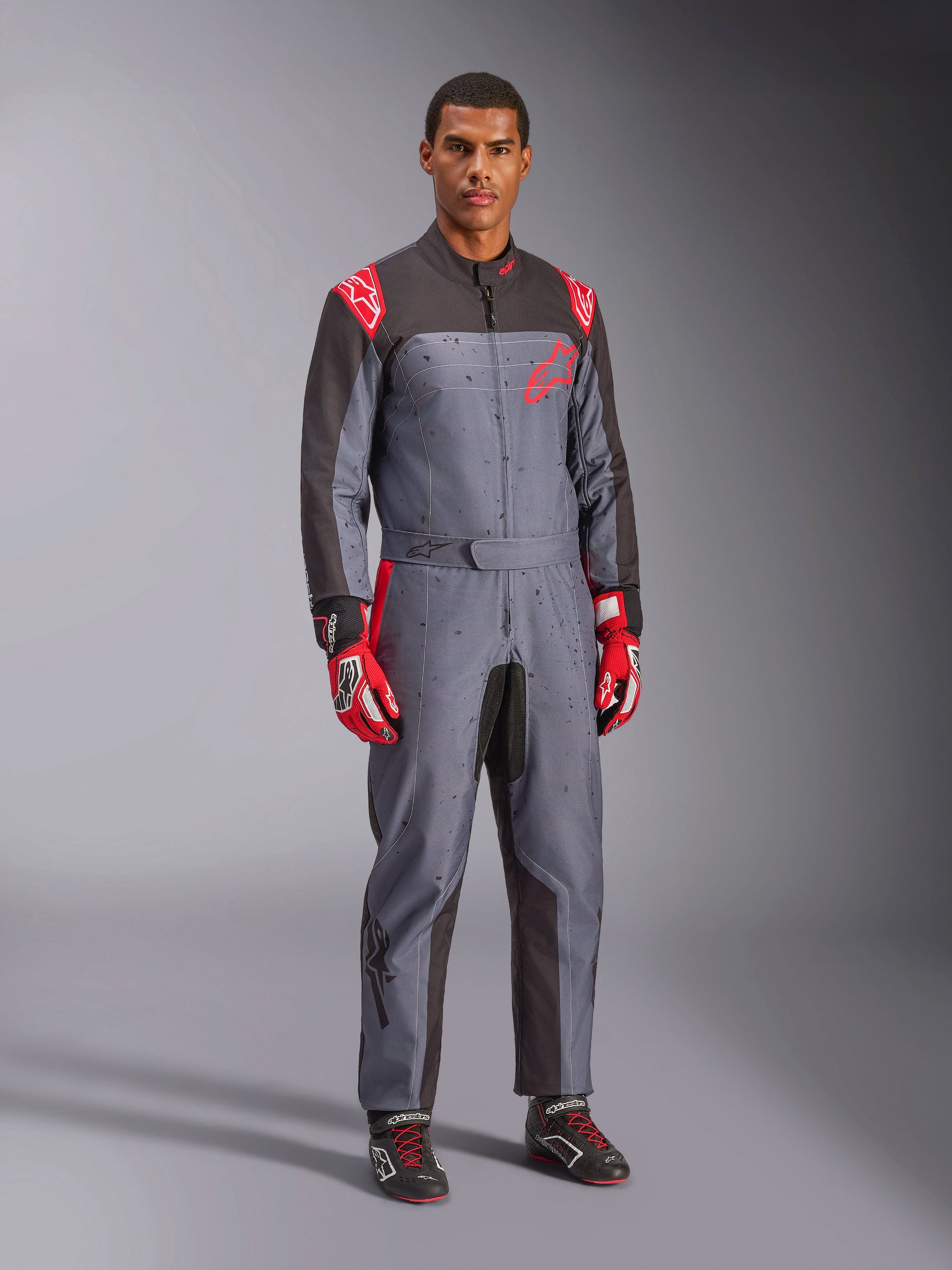 Tuta da karting Alpinestars KMX-9 V3 Graphic 6, grigio e antracite con dettagli rossi, caratterizzata da un'esclusiva grafica splash su petto e gambe, abbinata a guanti da corsa rossi e scarpe da karting nere, scatto a figura intera in studio.