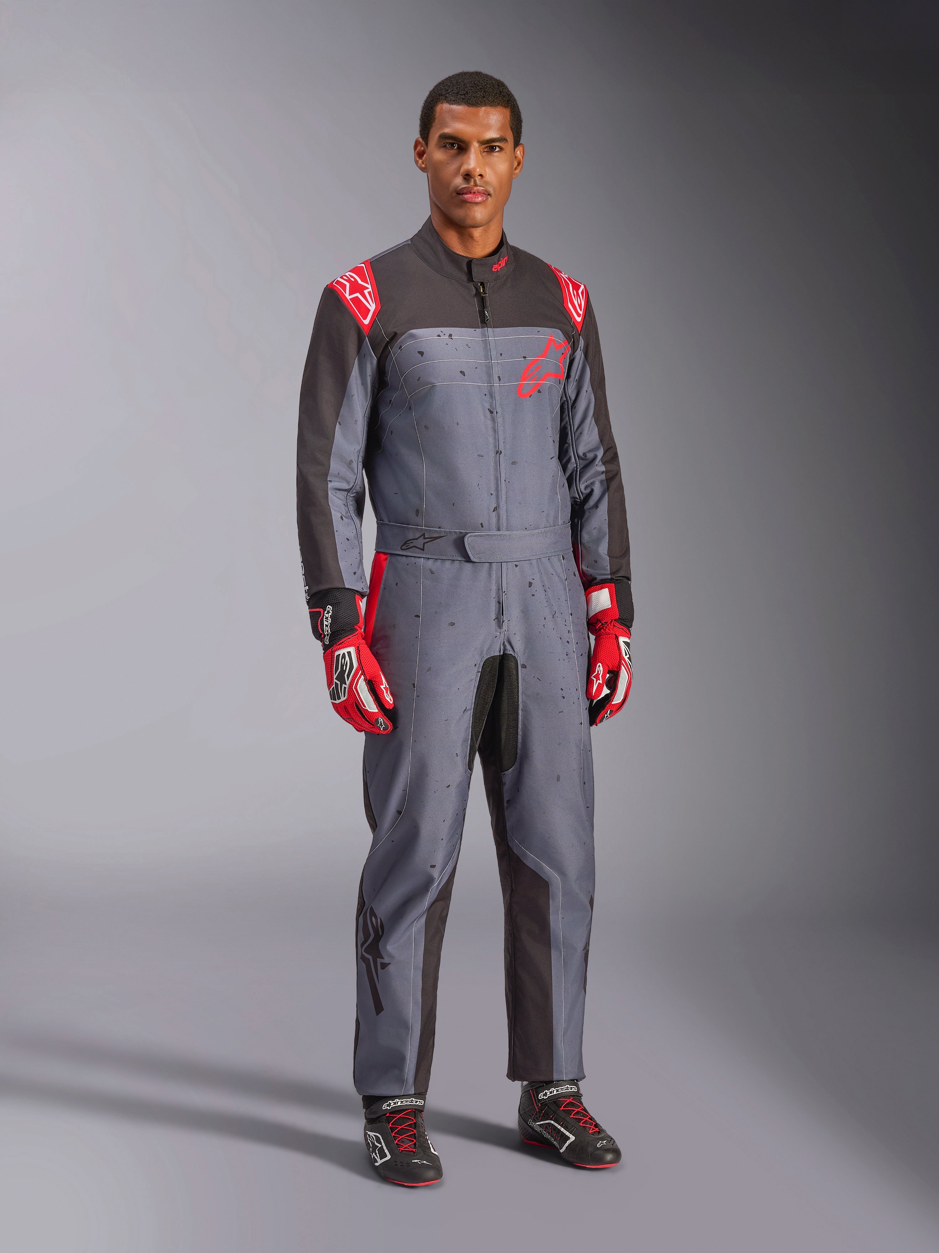 Tuta da karting Alpinestars KMX-9 V3 Graphic 6, grigio e antracite con dettagli rossi, caratterizzata da un'esclusiva grafica splash su petto e gambe, abbinata a guanti da corsa rossi e scarpe da karting nere, scatto a figura intera in studio.