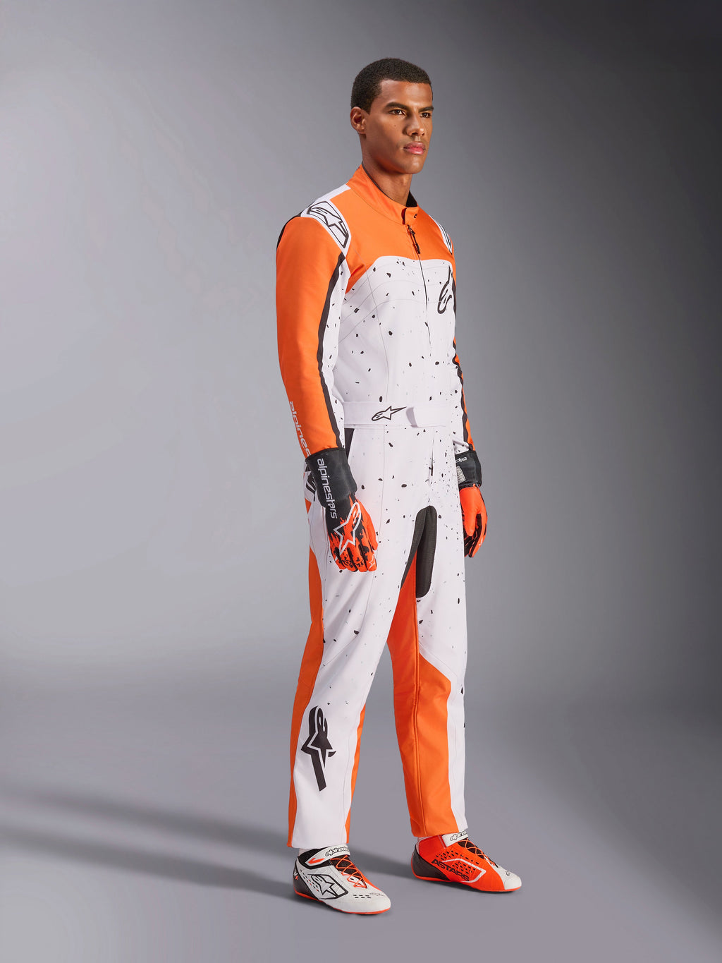Alpinestars KMX-9 V3 Graphic 6 Karting Suit, bianco, arancione e nero, con motivo punteggiato e vivaci maniche arancioni, indossata da un modello con guanti e scarpe da corsa coordinati, abbigliamento professionale da motorsport a doppio strato.