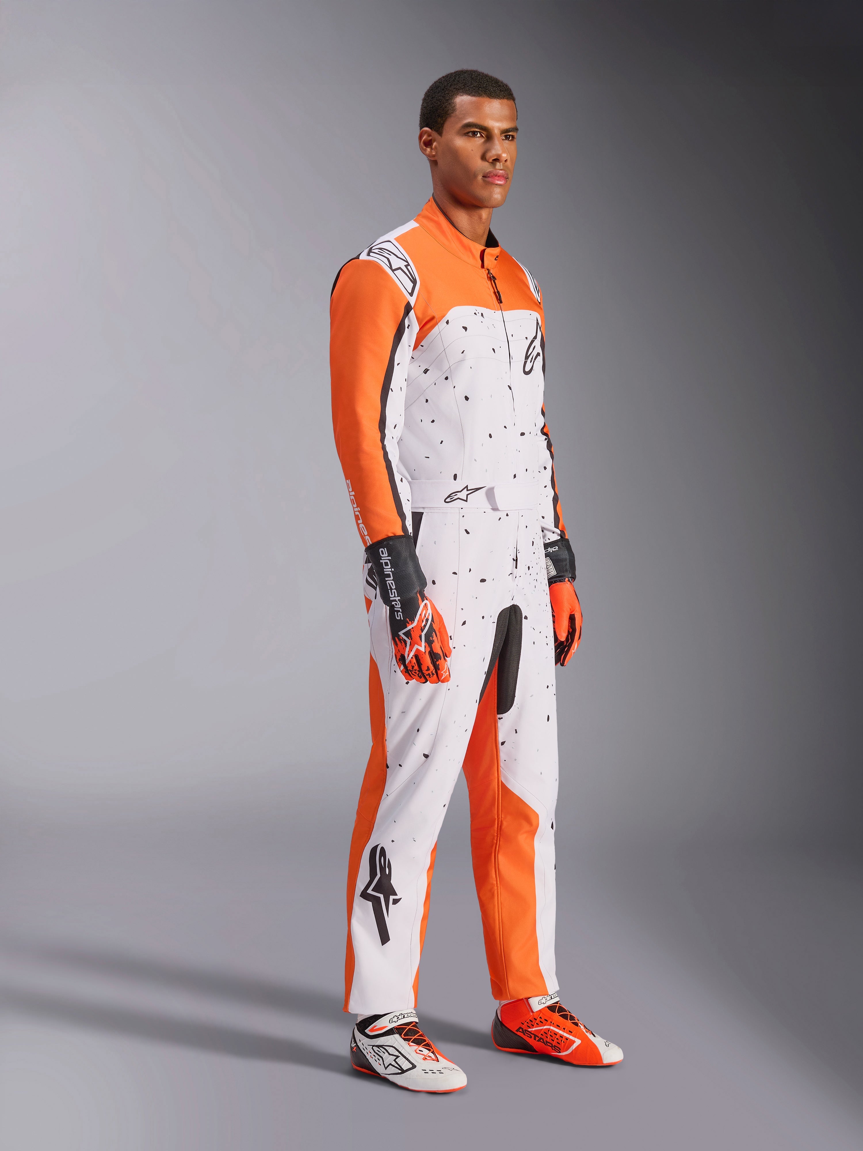 Alpinestars KMX-9 V3 Graphic 6 Karting Suit, bianco, arancione e nero, con motivo punteggiato e vivaci maniche arancioni, indossata da un modello con guanti e scarpe da corsa coordinati, abbigliamento professionale da motorsport a doppio strato.