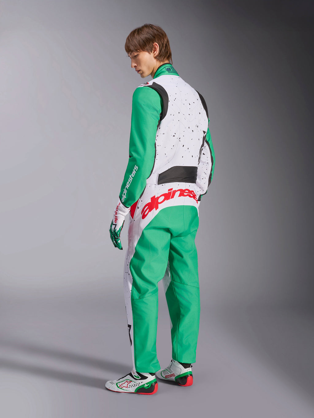Tuta Alpinestars KMX-9 V3 Graphic 6, tuta da karting, bianco, verde e rosso, modello visto dal retro, con maniche e pannelli inferiori verdi, parte superiore del busto bianca con dettagli a macchioline nere, vistoso marchio Alpinestars rosso sulla parte bassa della schiena, abbinata a scarpe da karting bianche e verdi coordinate.