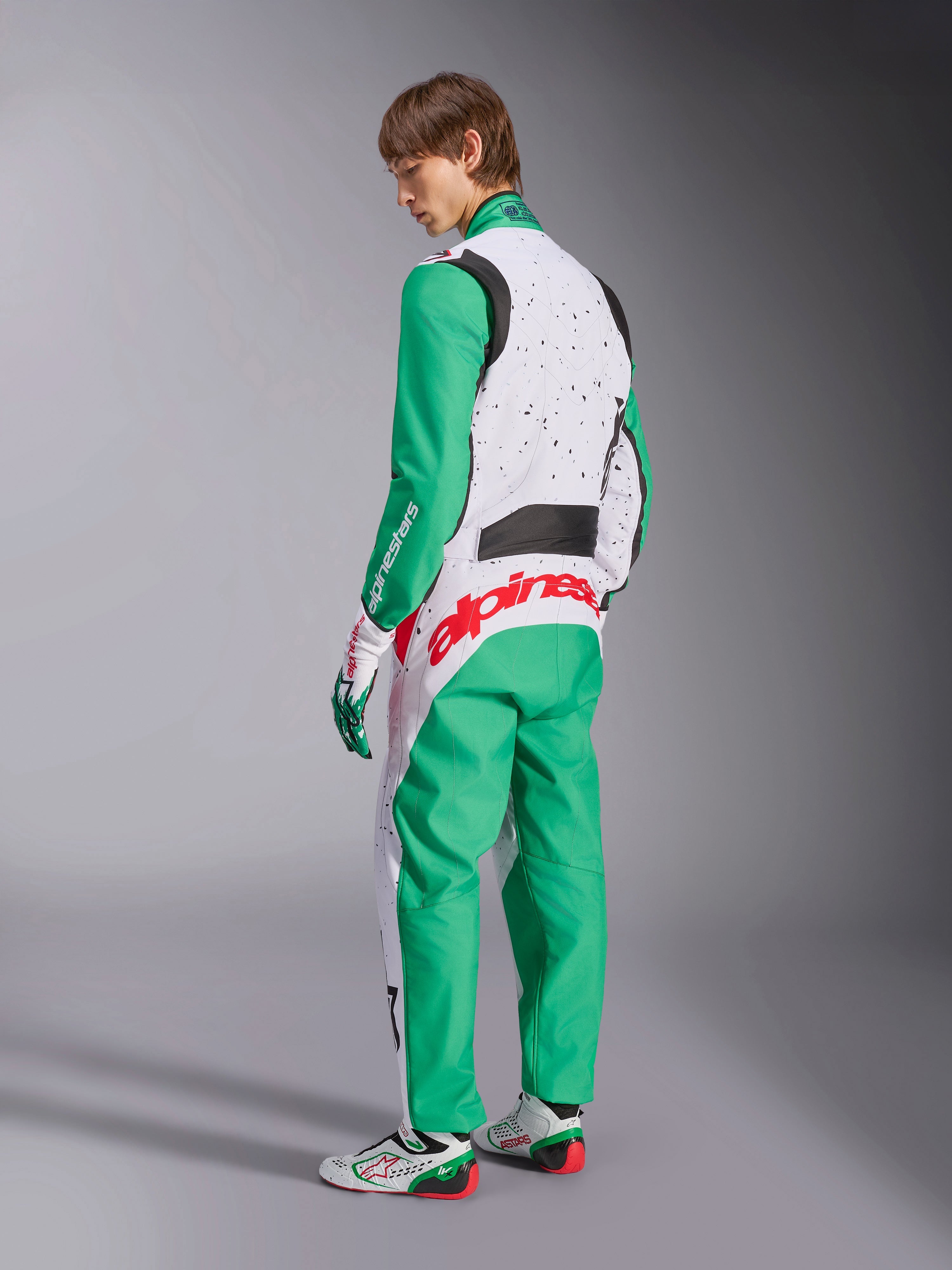 Tuta Alpinestars KMX-9 V3 Graphic 6, tuta da karting, bianco, verde e rosso, modello visto dal retro, con maniche e pannelli inferiori verdi, parte superiore del busto bianca con dettagli a macchioline nere, vistoso marchio Alpinestars rosso sulla parte bassa della schiena, abbinata a scarpe da karting bianche e verdi coordinate.
