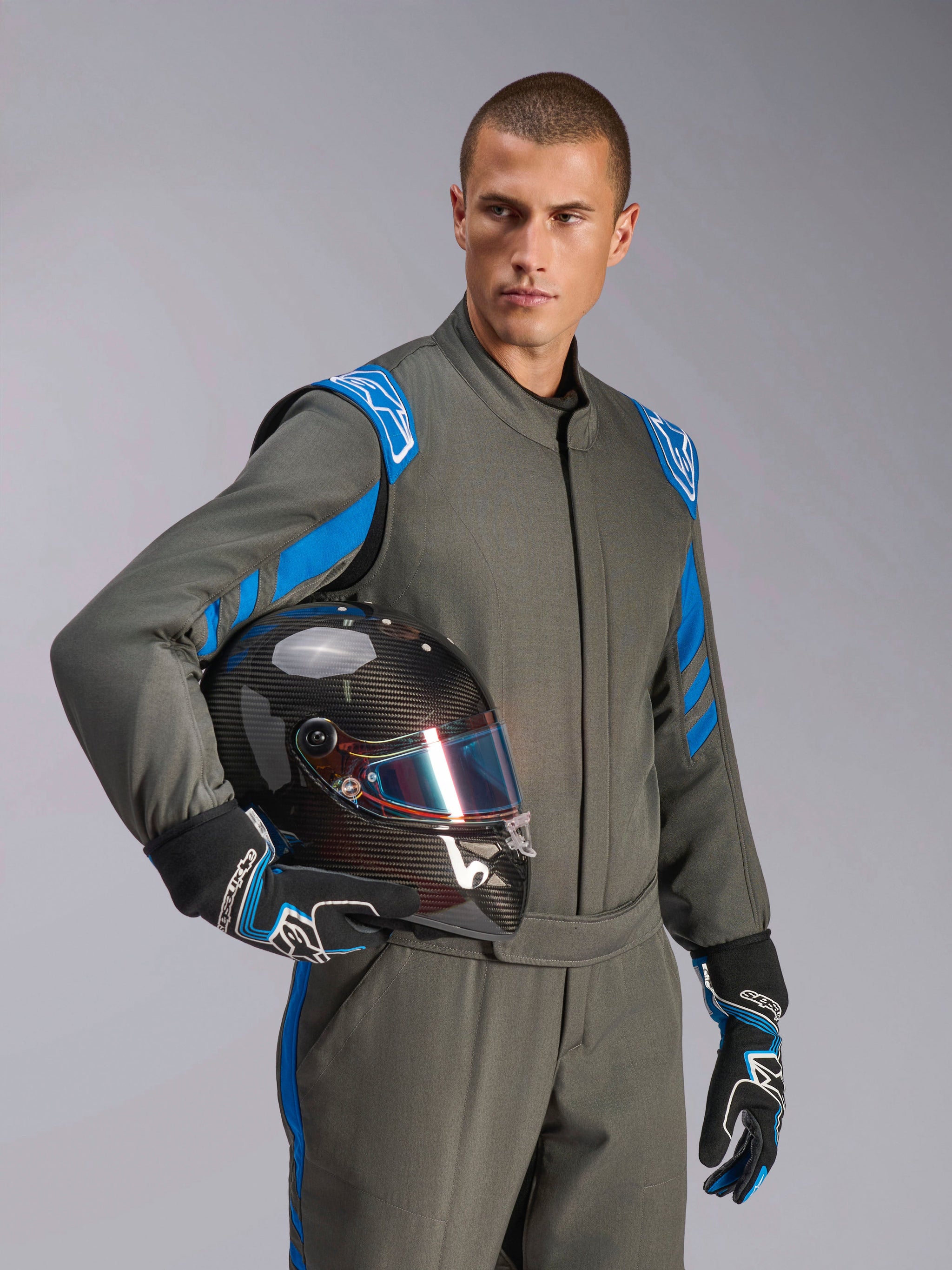 Pilota Alpinestars con tuta GP Race V3 FIA in Anthracite Blue, casco da corsa in fibra di carbonio con visiera iridescente, abbigliamento professionale per corse auto con dettagli blu sulle spalle e guanti coordinati.