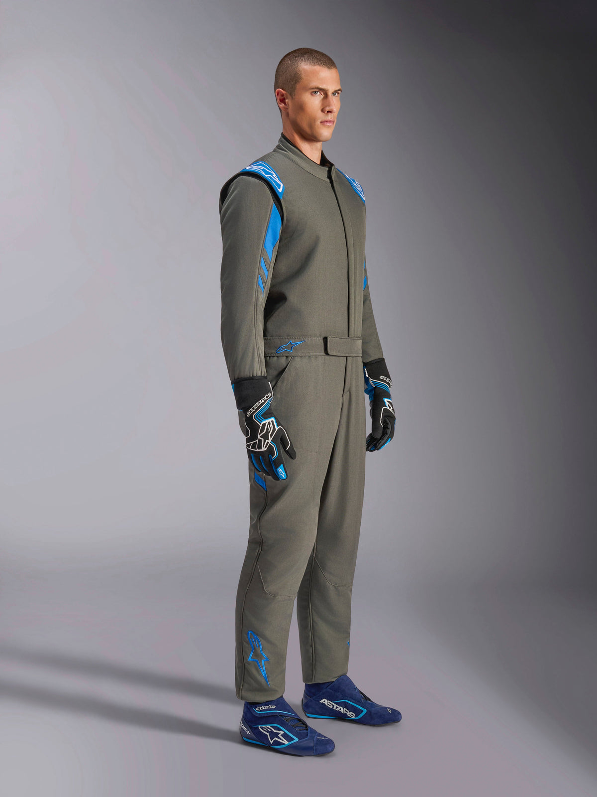 Tuta Alpinestars GP Race V3 FIA in Antracite Blu, tuta intera da corsa indossata da un modello, caratterizzata da accenti blu brillante su spalle e caviglie, abbinata a guanti da corsa coordinati e scarpe da corsa blu, equipaggiamento professionale per motorsport.